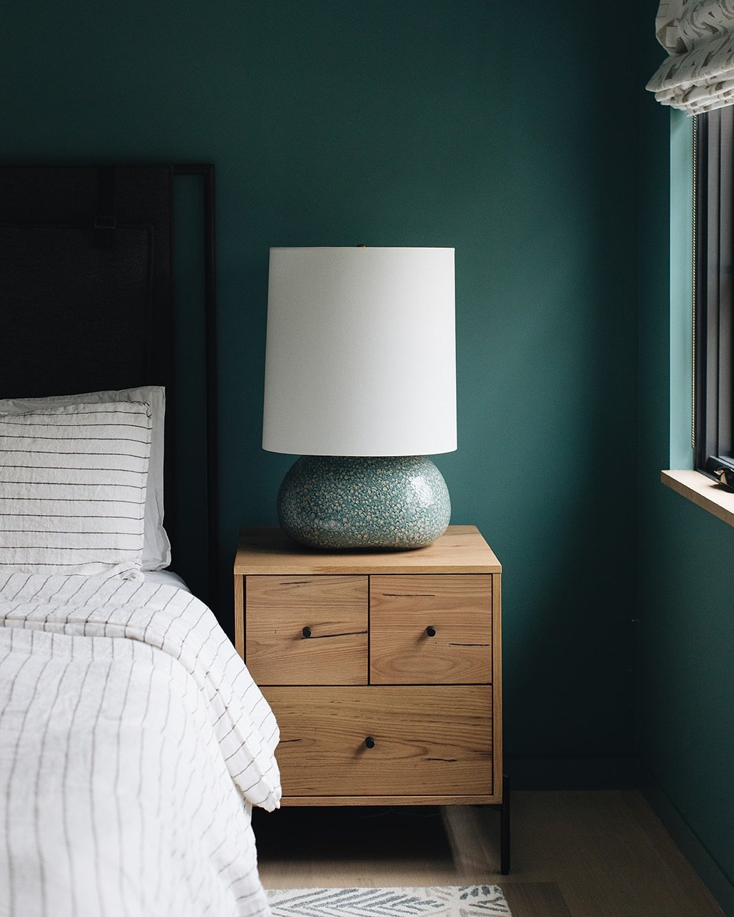A light oak nightstand with two asymmetrical drawers and black pulls. On top is a large, textured blue-green ceramic lamp with a white drum shade. The bedroom wall is painted a rich teal color.