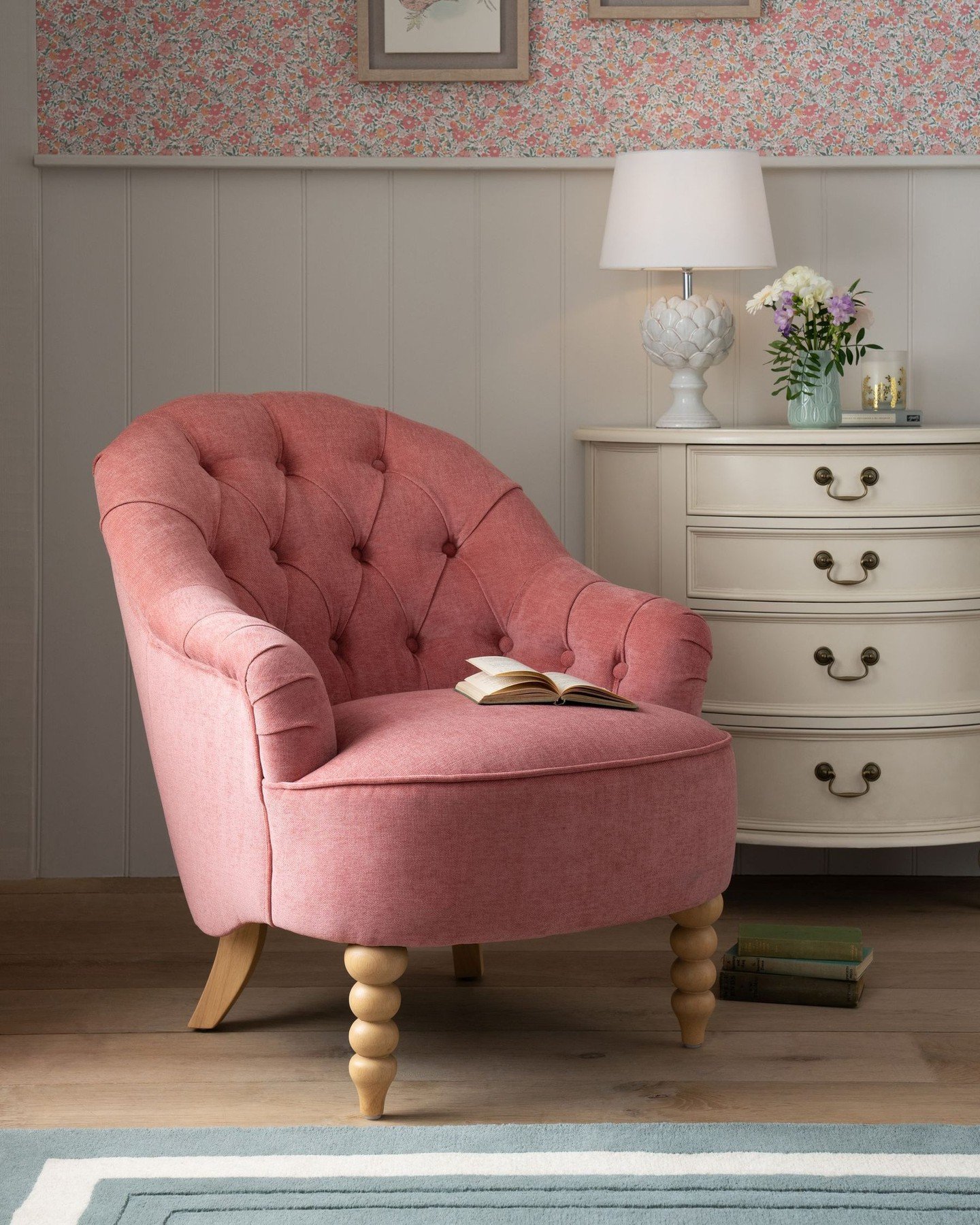 A tufted coral-pink armchair with natural turned wood legs sits on a wooden floor in a bedroom.