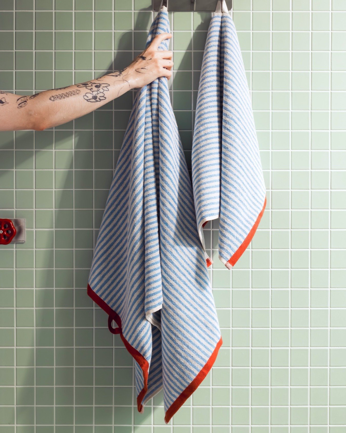 Two blue and white diagonal striped towels with a coral-red border hanging on a hook against mint green tile.