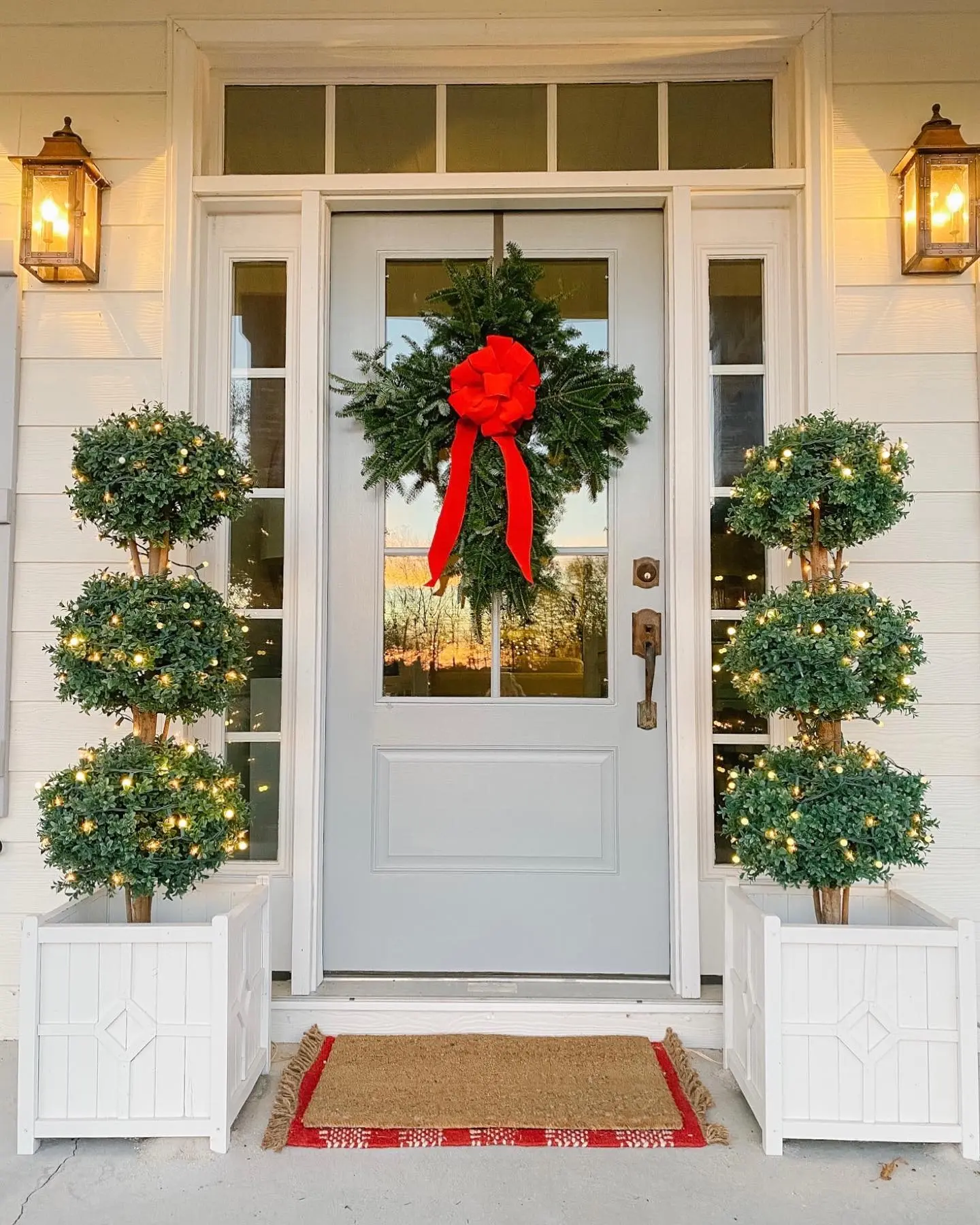 A bright Christmas porch with a light gray door, flanked by two illuminated three-tier boxwood topiaries in white wooden planters, and a simple evergreen cross-wreath with a large red bow.
