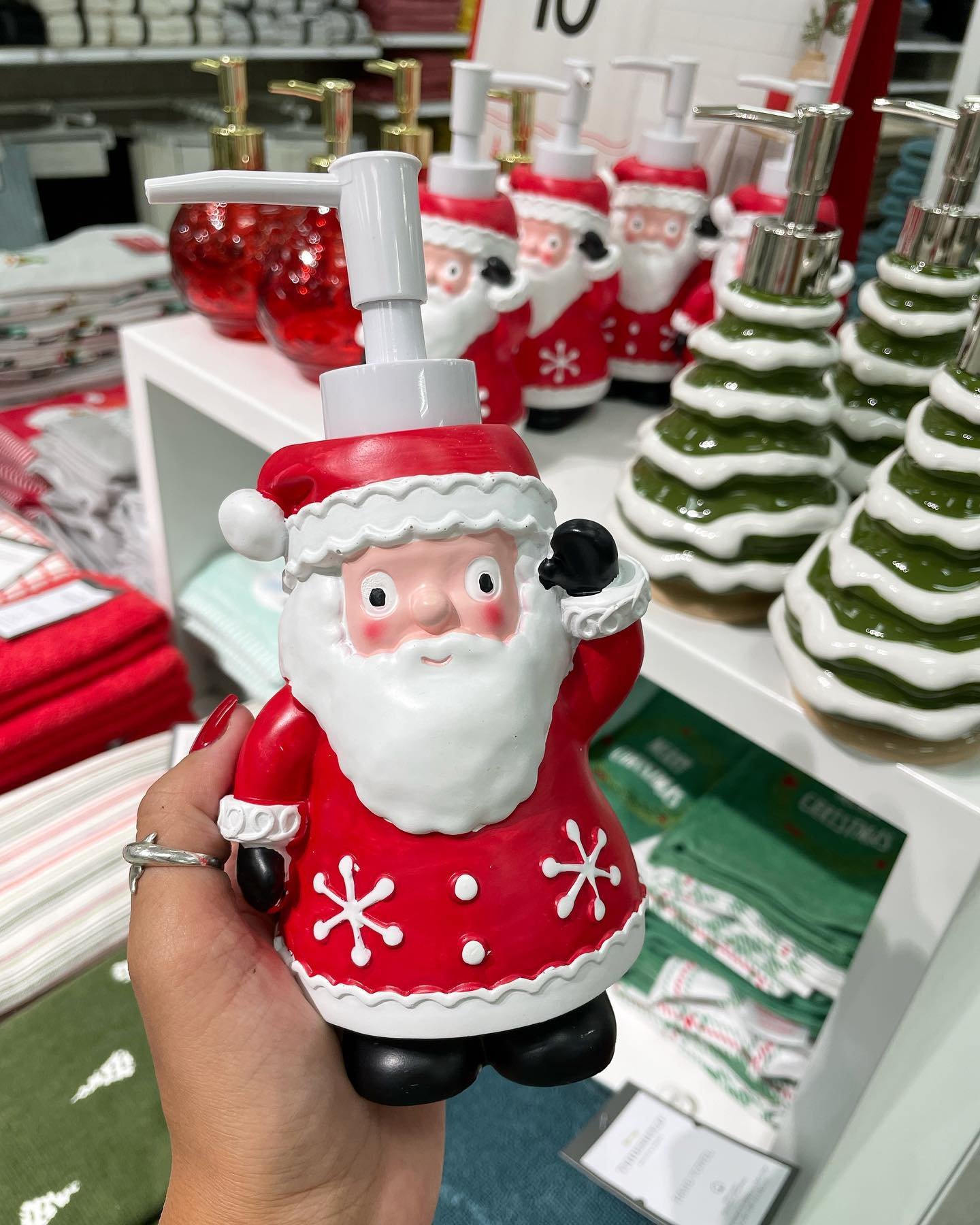 A hand holding a ceramic soap dispenser shaped like Santa Claus in a retail store with other Christmas tree dispensers in the background.