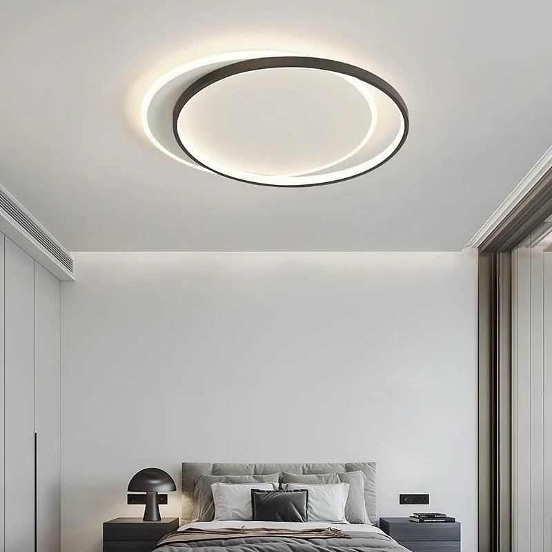 A modern, minimalist flush mount ceiling light featuring two interlocking black and white rings sits against a white ceiling over a bed.