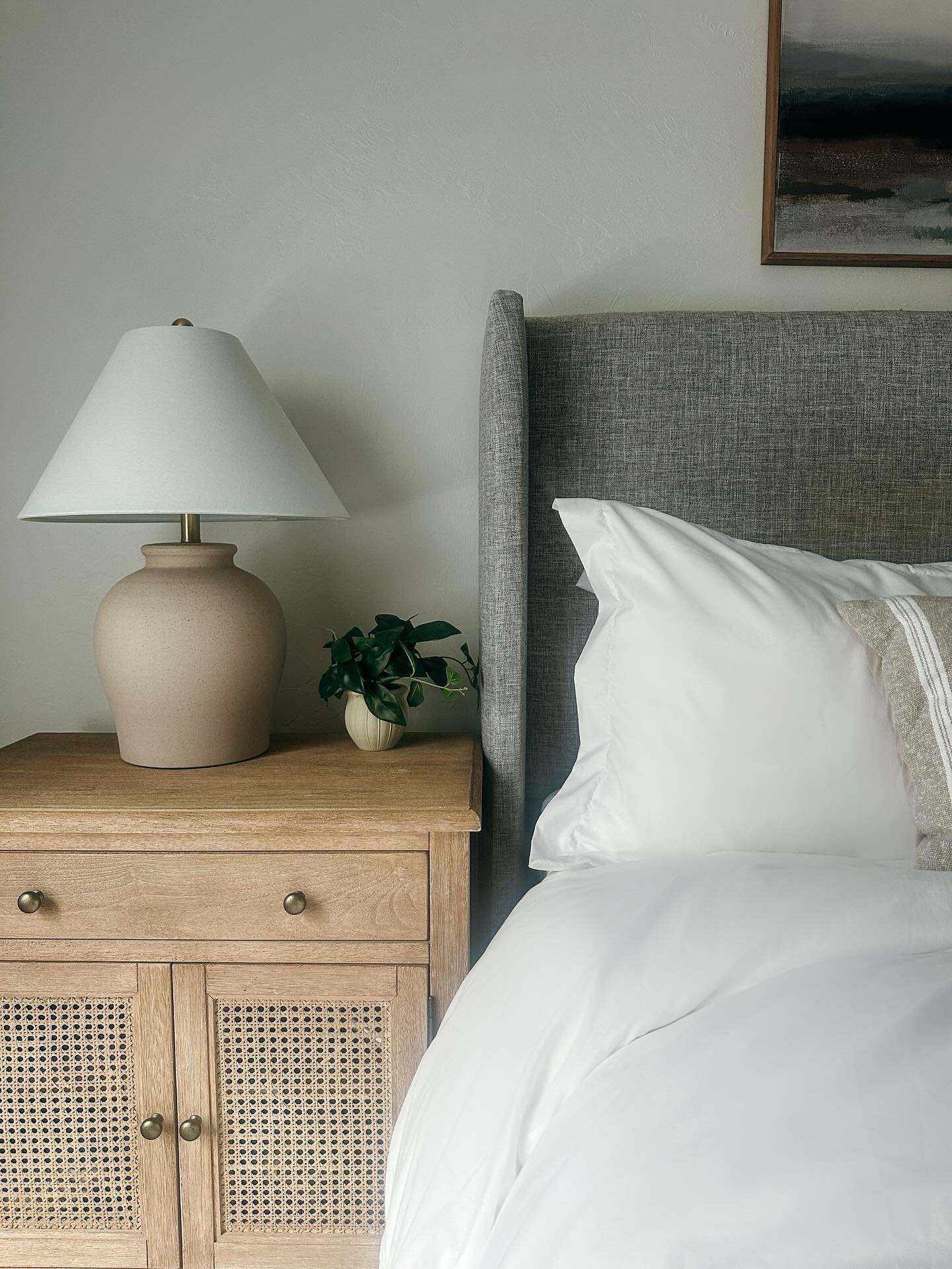 A neutral-toned bedroom with a textured, rounded lamp sitting on a wooden nightstand with rattan cabinet doors.