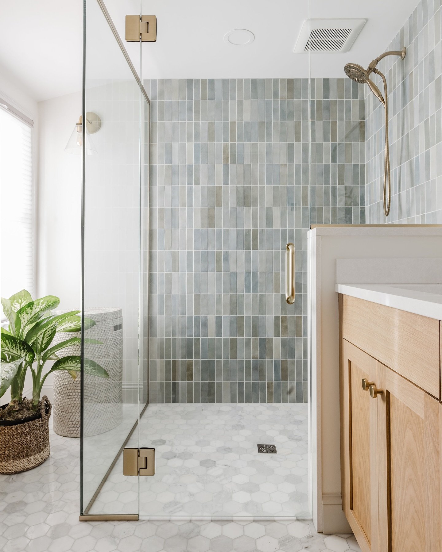 A modern walk-in shower with a mosaic of blue, gray, and white vertical tiles on the back wall, a white hexagonal mosaic floor, and light wood vanity.