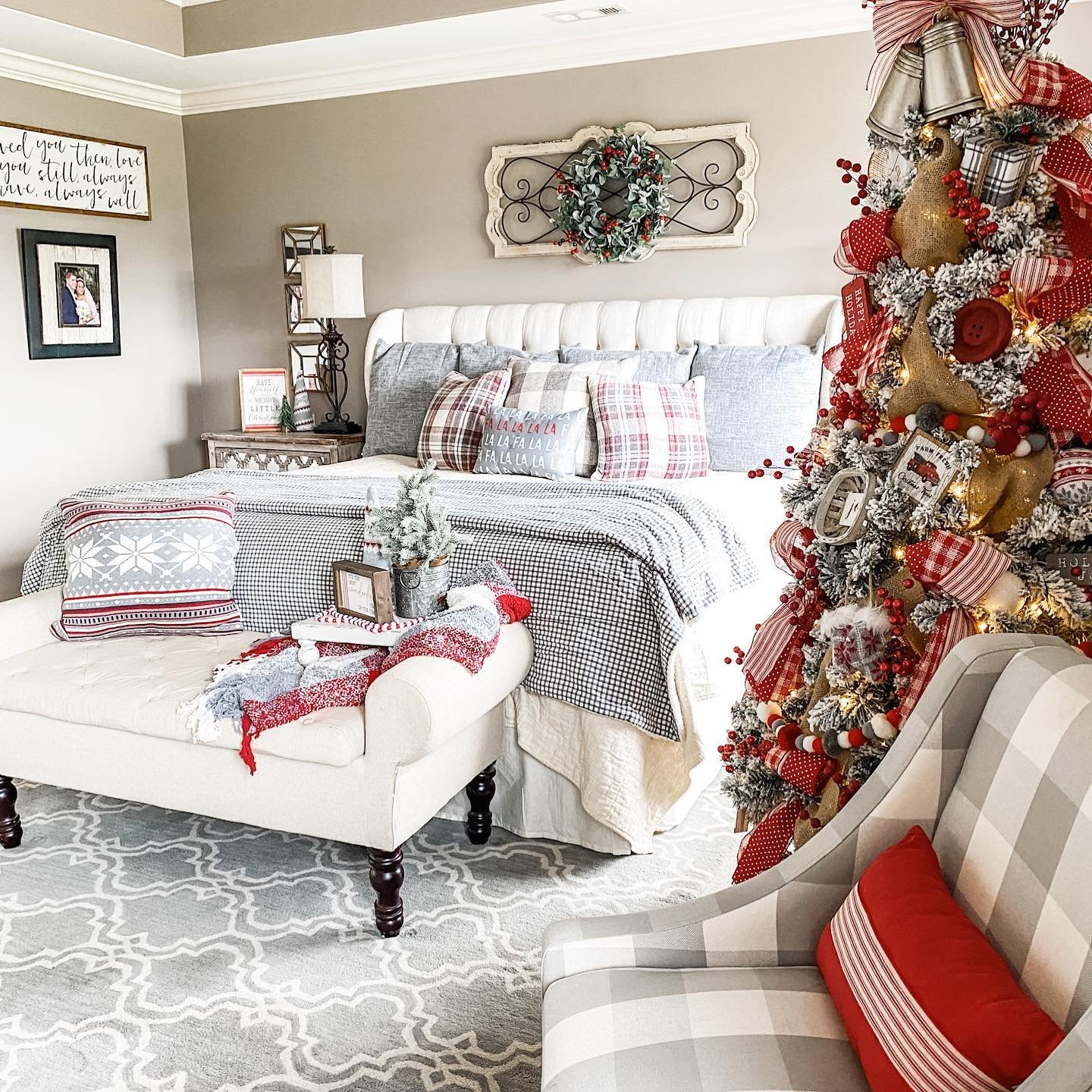 A neutral-toned farmhouse bedroom featuring a bed dressed in gray and white buffalo check, red and gray plaid pillows, a large, heavily decorated Christmas tree, and a matching plaid accent chair.