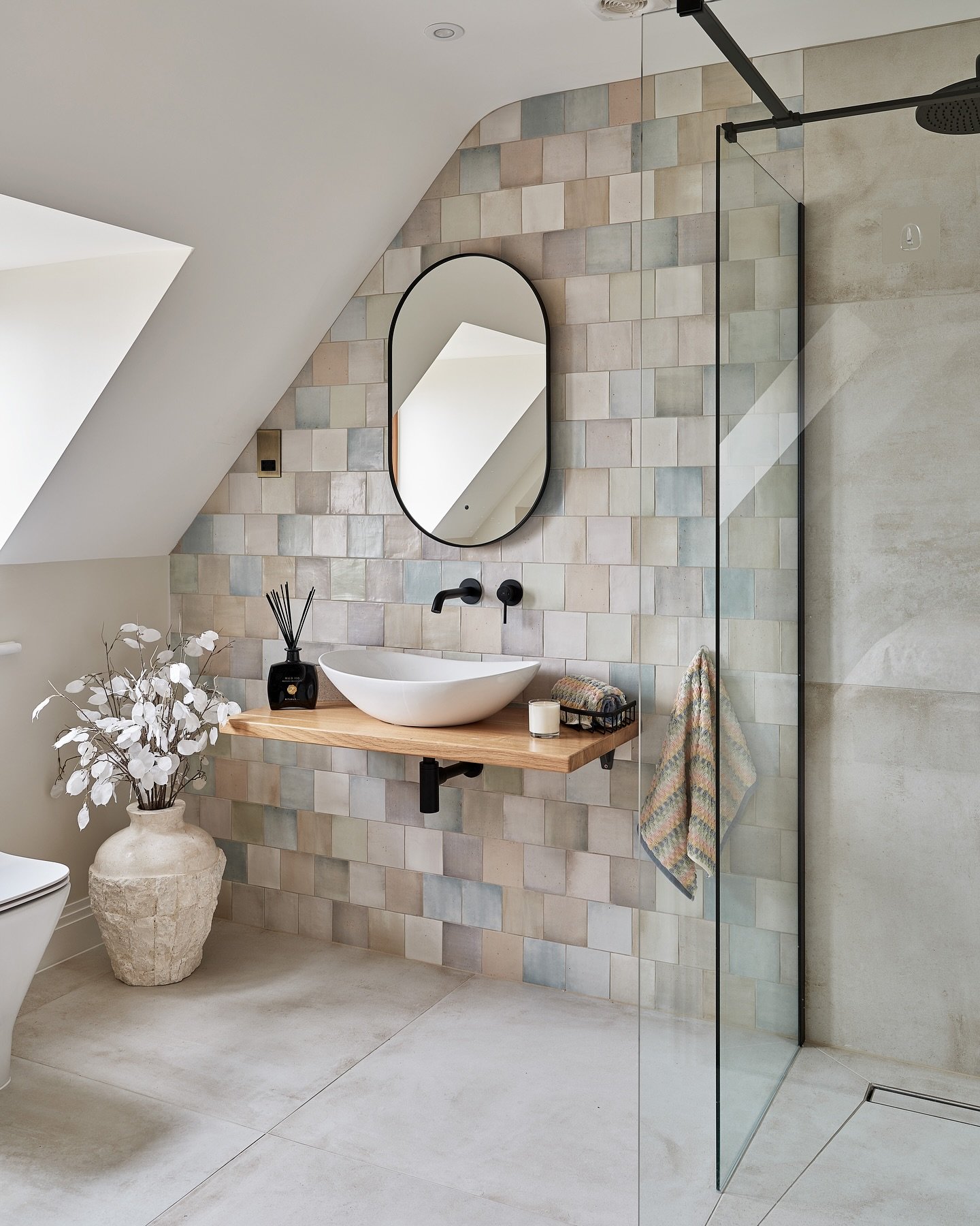 A modern bathroom with a sloped ceiling, a floating wood shelf vanity, and a feature wall tiled with a patchwork of square tiles in varying pastel shades of blue, beige, and white.