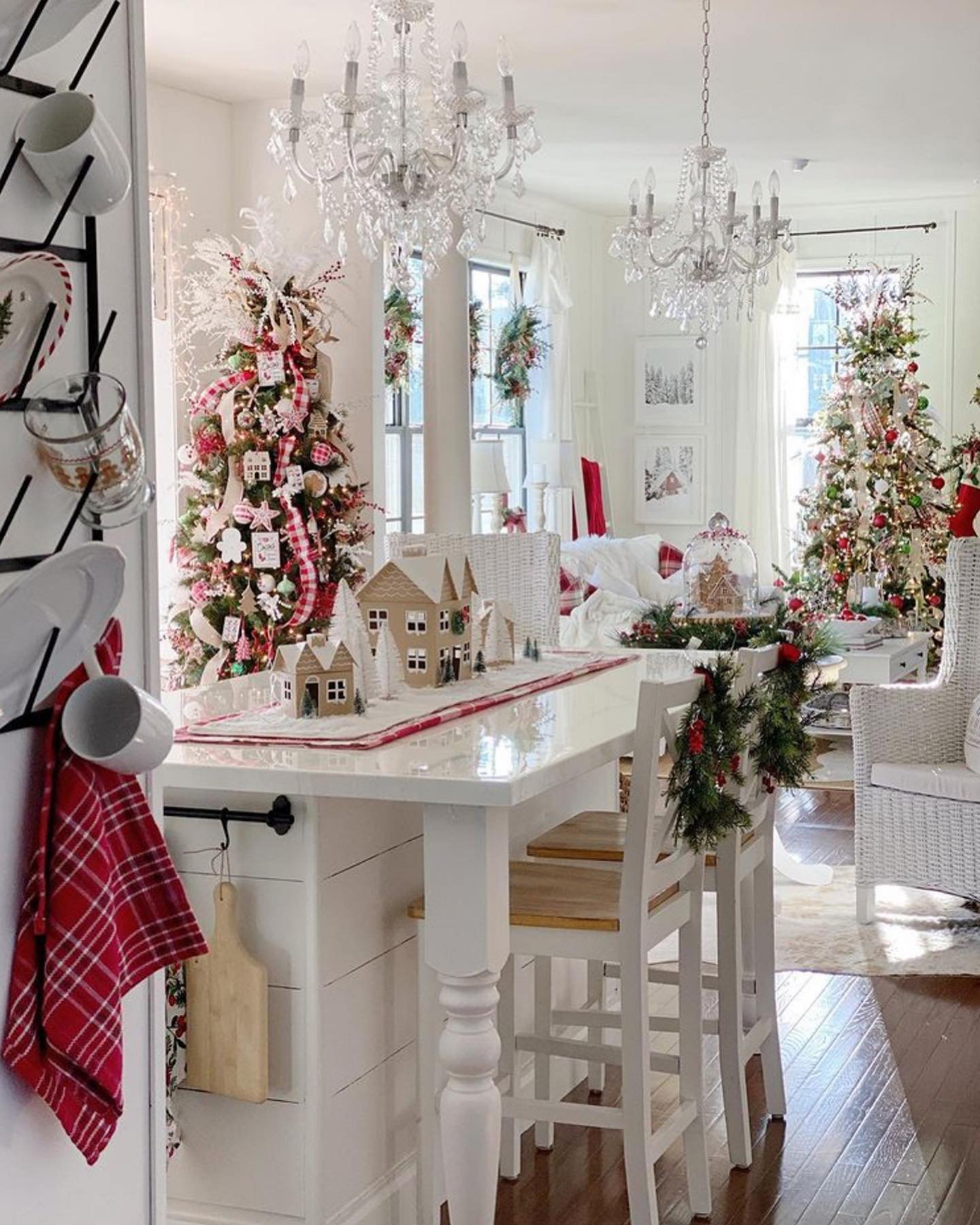 A bright, high-end farmhouse kitchen and dining area featuring two large, heavily decorated Christmas trees, white shiplap island with red and white gingham accents, and crystal chandeliers overhead.