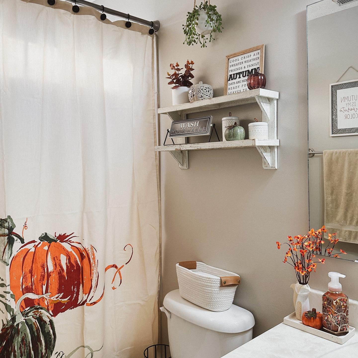 A neutral-toned bathroom with a shower curtain featuring a large pumpkin illustration and a rustic shelf with fall decorations.