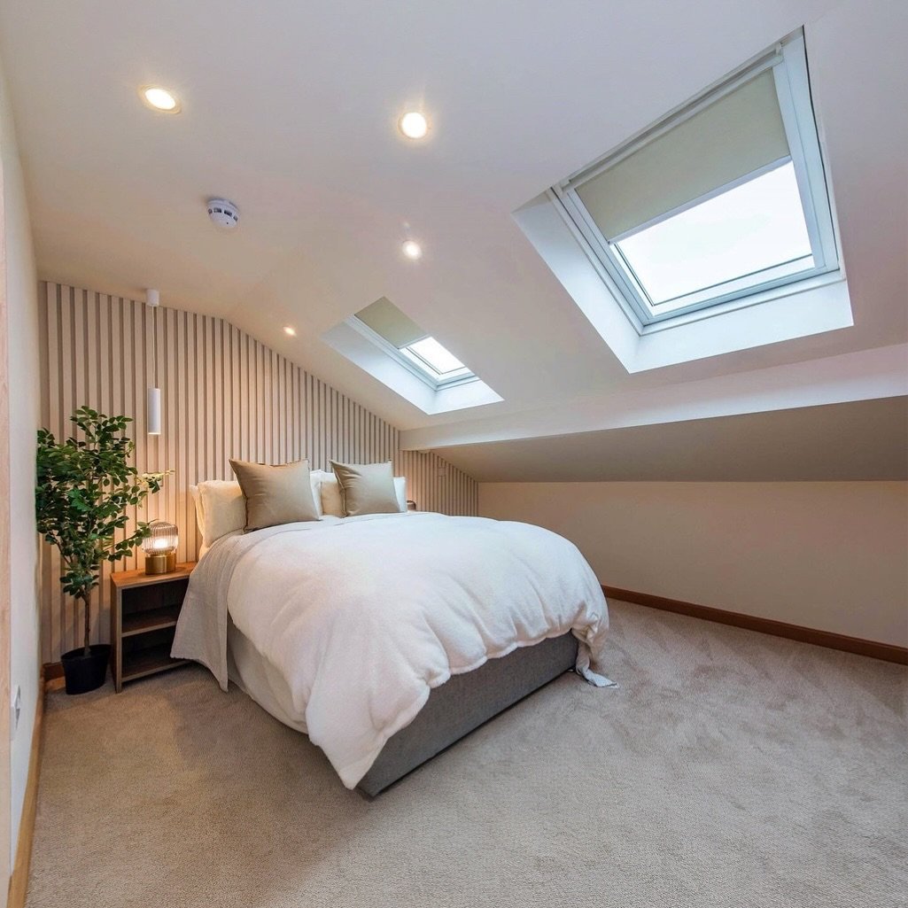 A bright loft bedroom with sloped ceilings and multiple skylights. The room has a wood-slatted accent wall behind a low gray and white bed.
