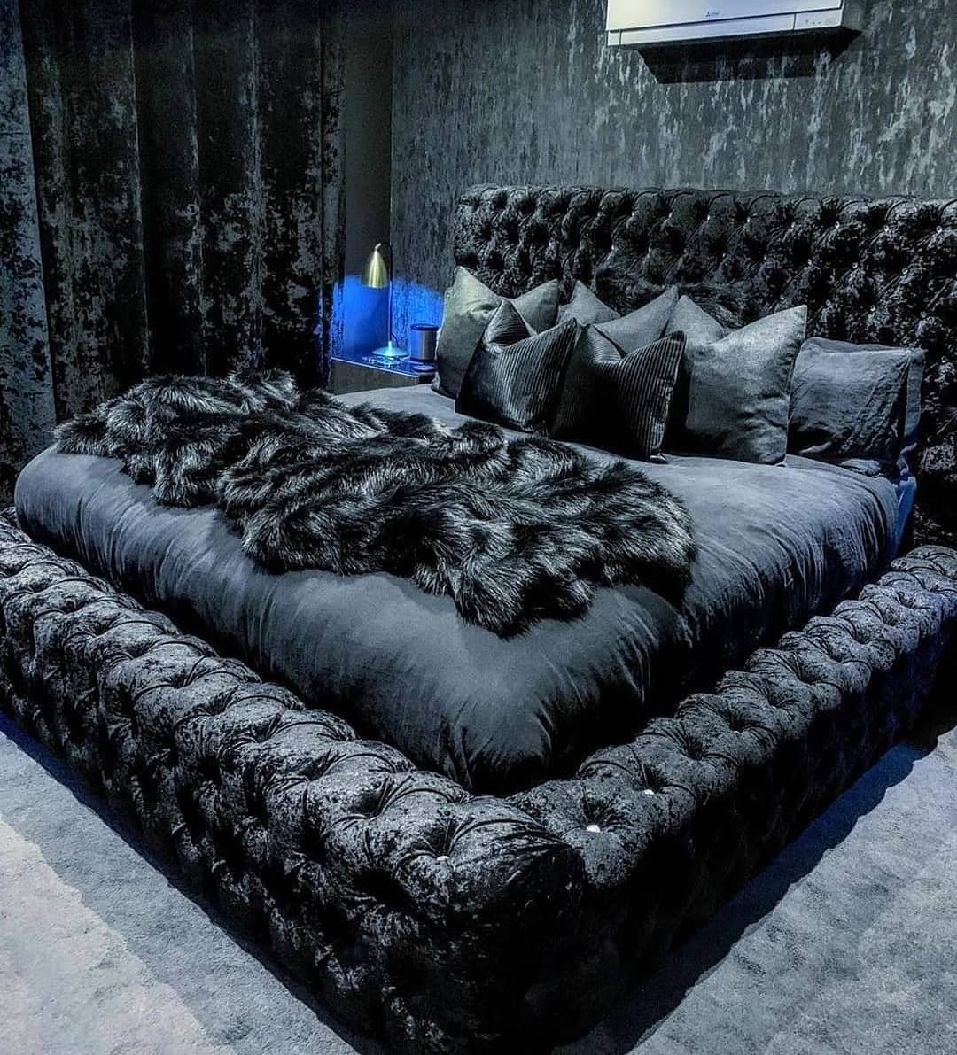 A luxurious and moody bedroom featuring a large tufted black velvet bed with faux fur blankets and dark, textured wallpaper.