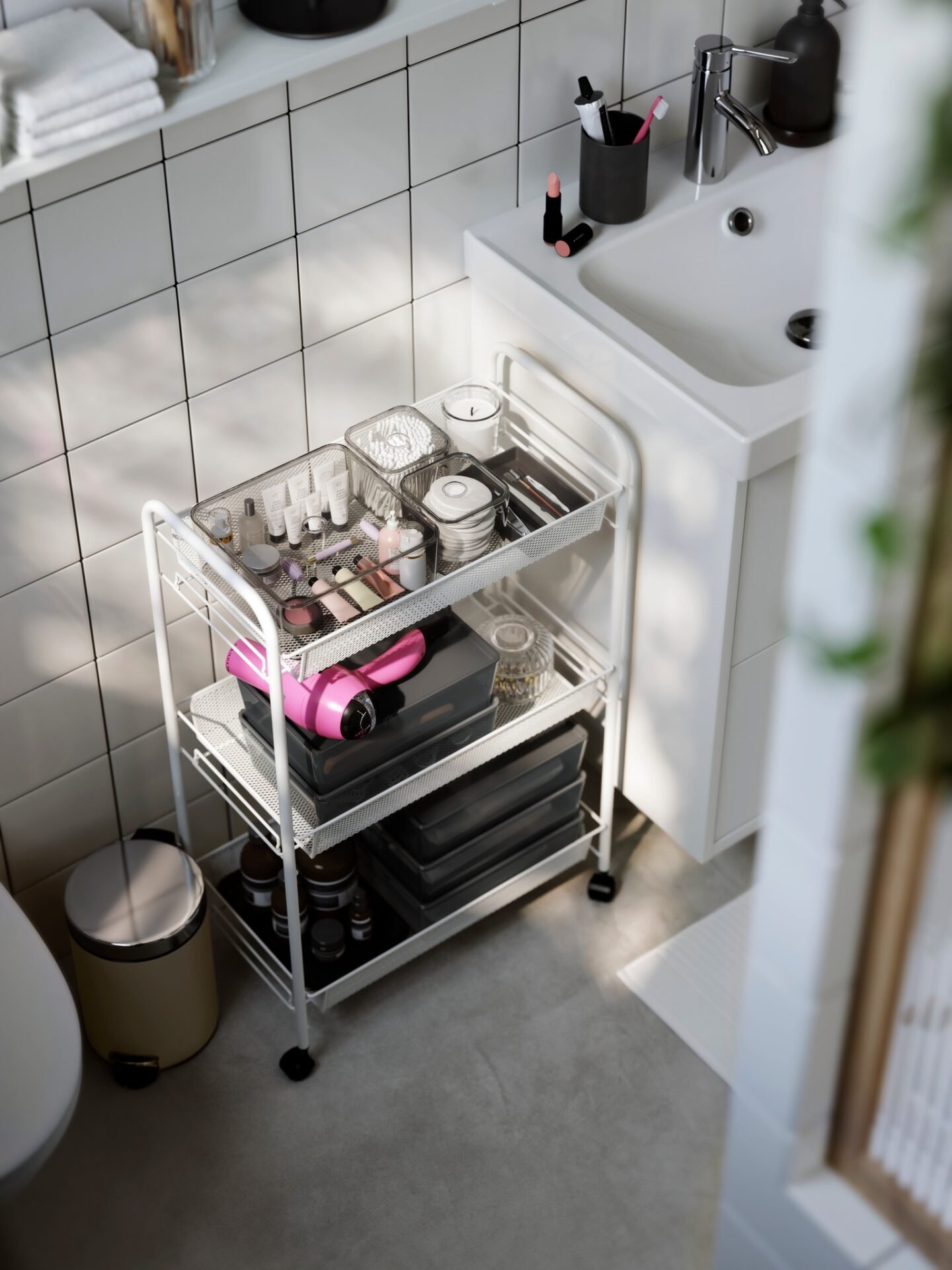 White metal three-tier rolling cart positioned next to a pedestal sink, organized with makeup, toiletries, a hairdryer, and storage boxes.