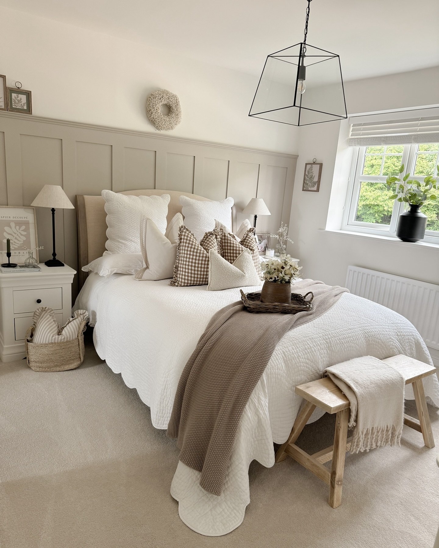 A black metal lantern-style pendant light with a clear glass shade hangs over a bed in a cozy, neutral-toned bedroom.