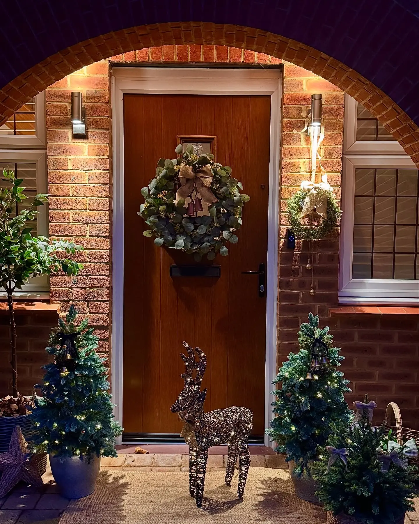 A warmly lit Christmas porch at night featuring two small, potted evergreen trees, a sparkling wire reindeer, and a large wreath made of eucalyptus leaves on a wooden door framed by brick archway.
