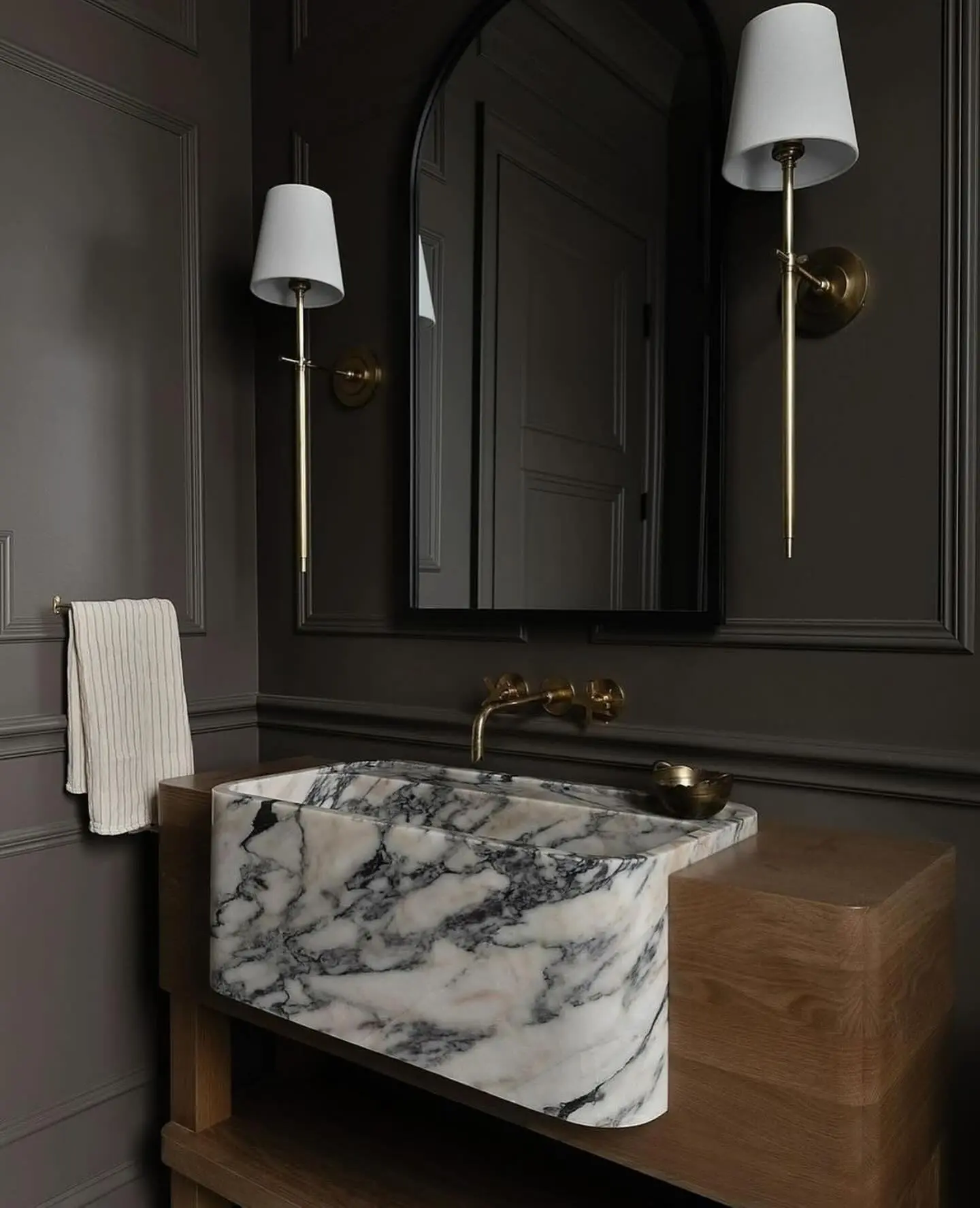 A dark bathroom with a unique vanity featuring a white and gray veined marble basin that drapes over the front edge, resting on a light wood cabinet base. A black arched mirror and two long brass sconces hang above it, with a wall-mounted brass faucet.