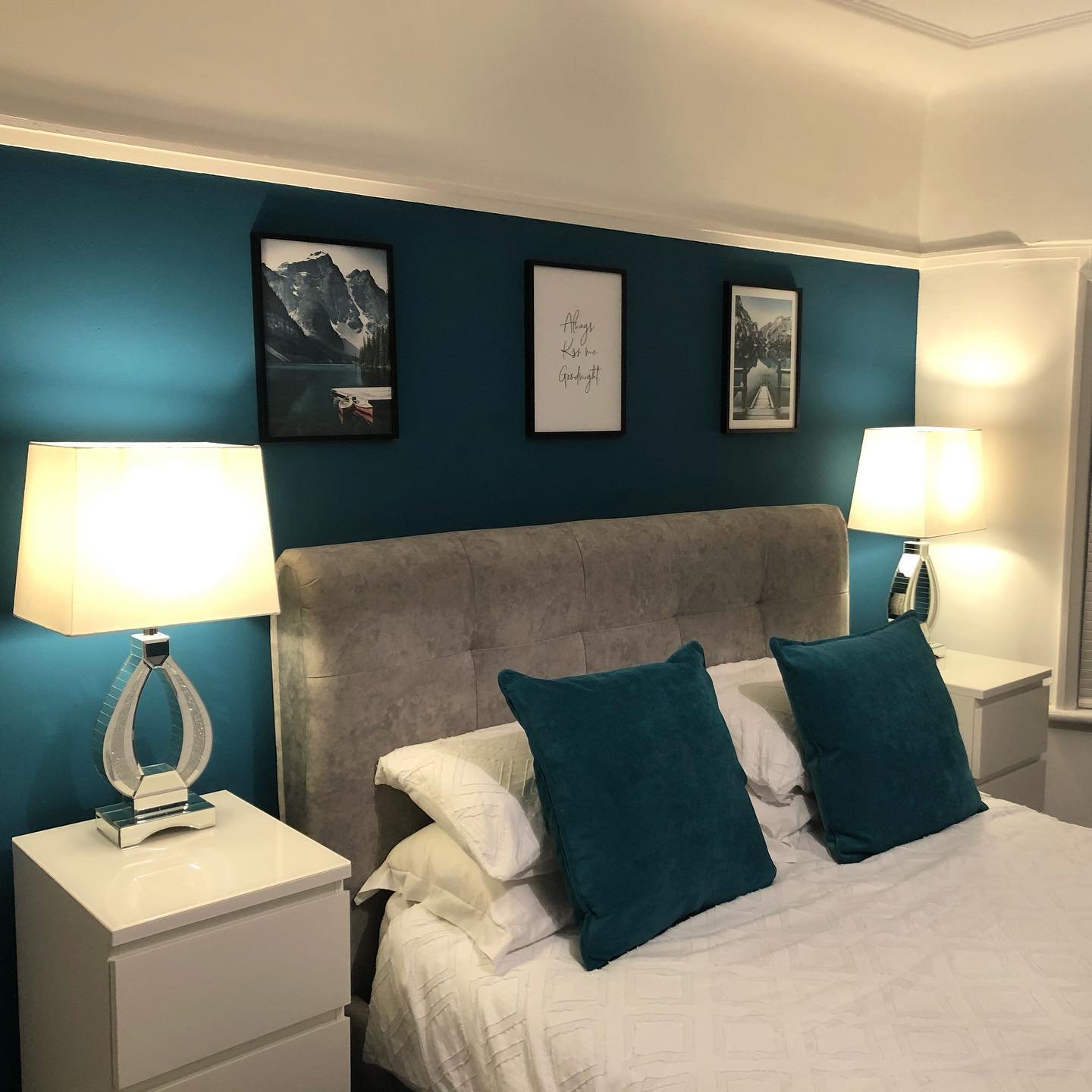Two modern, chrome-based lamps with white shades flanking a bed with a tufted headboard against a teal accent wall.