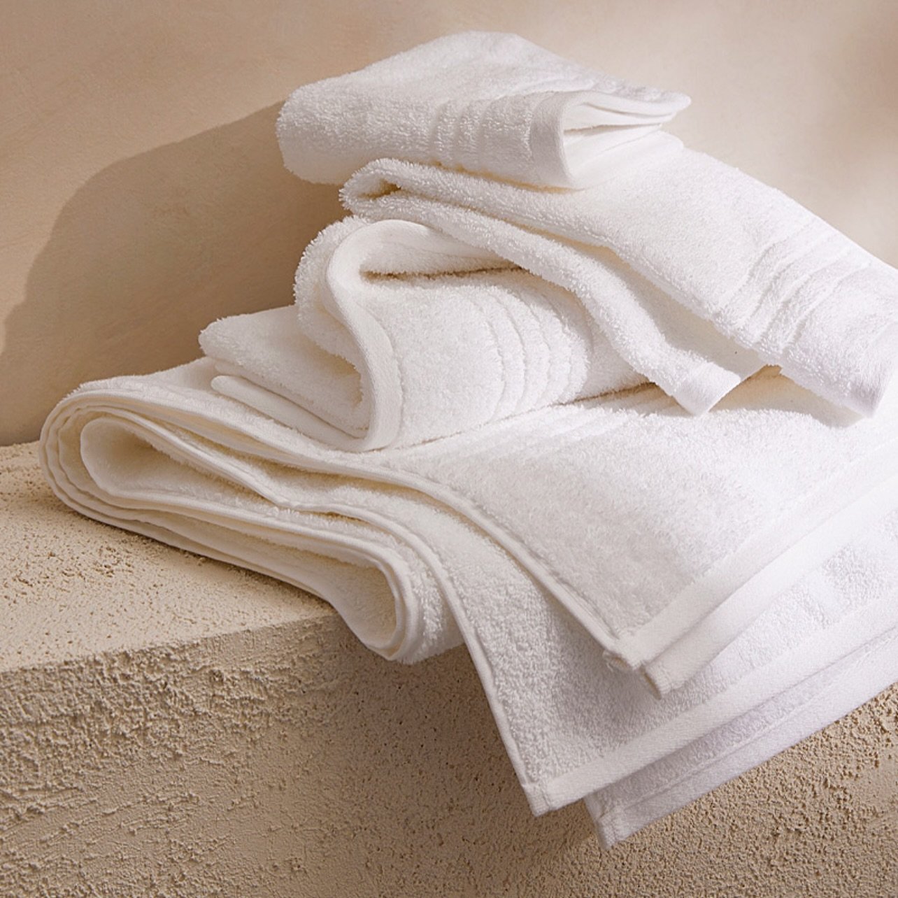 A casually stacked collection of pure white, plush terry cloth towels on a textured beige stone surface.