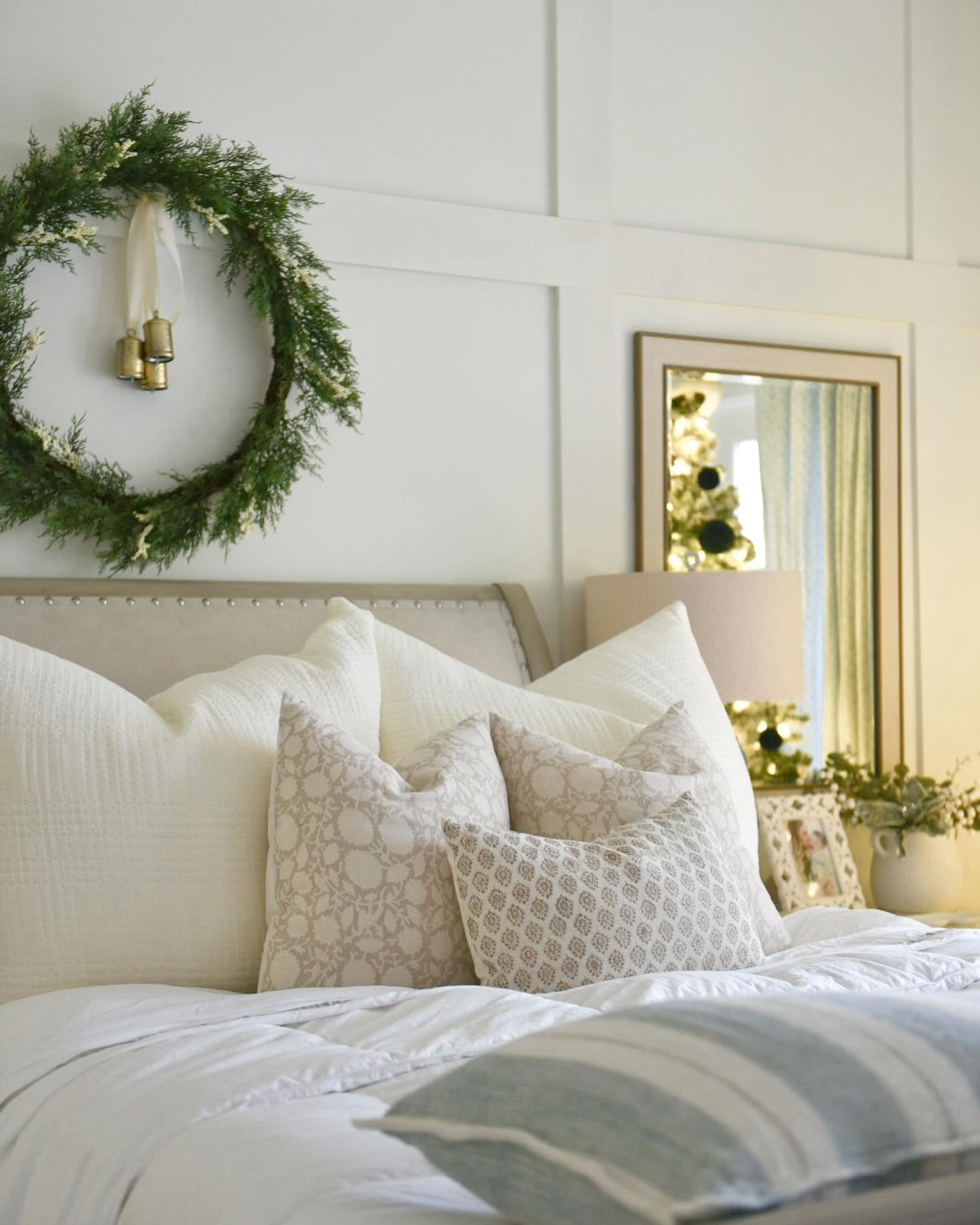  A close-up of a bright, serene bedroom featuring all-white bedding, neutral patterned throw pillows, and a simple green cedar wreath hanging above the bed with two brass bells.