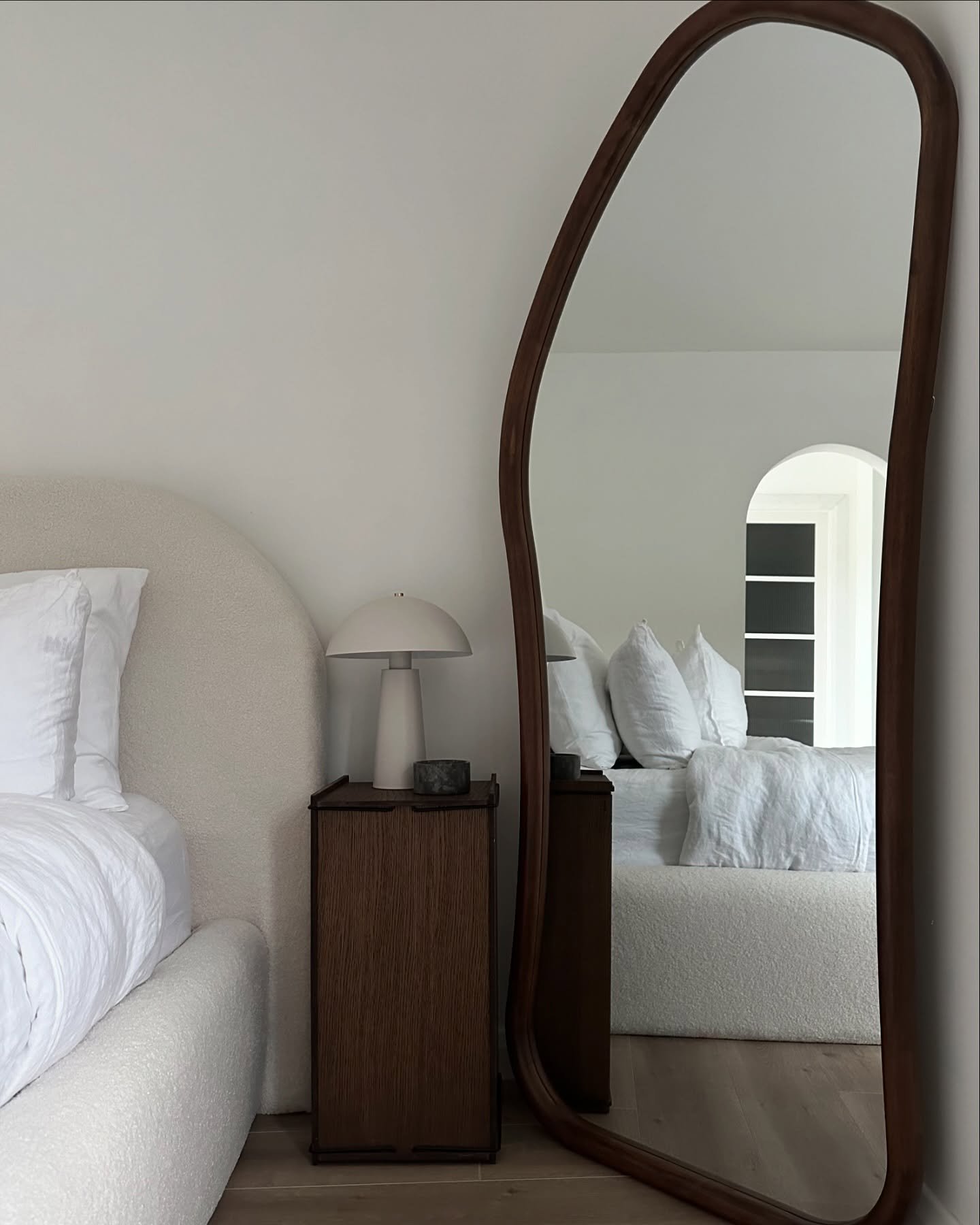 A tall, dark wood-framed mirror with an irregular, wavy shape leans in a modern bedroom, reflecting a bed with a white headboard and a white lamp on a nightstand.