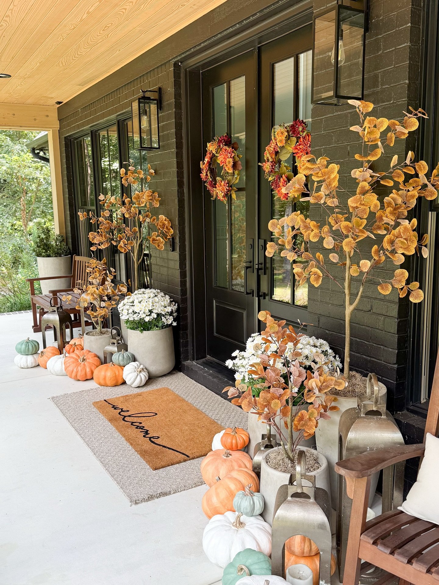 A stylish, modern farmhouse porch with black brick walls, decorated with pumpkins in various colors and concrete planters with trees and white mums.