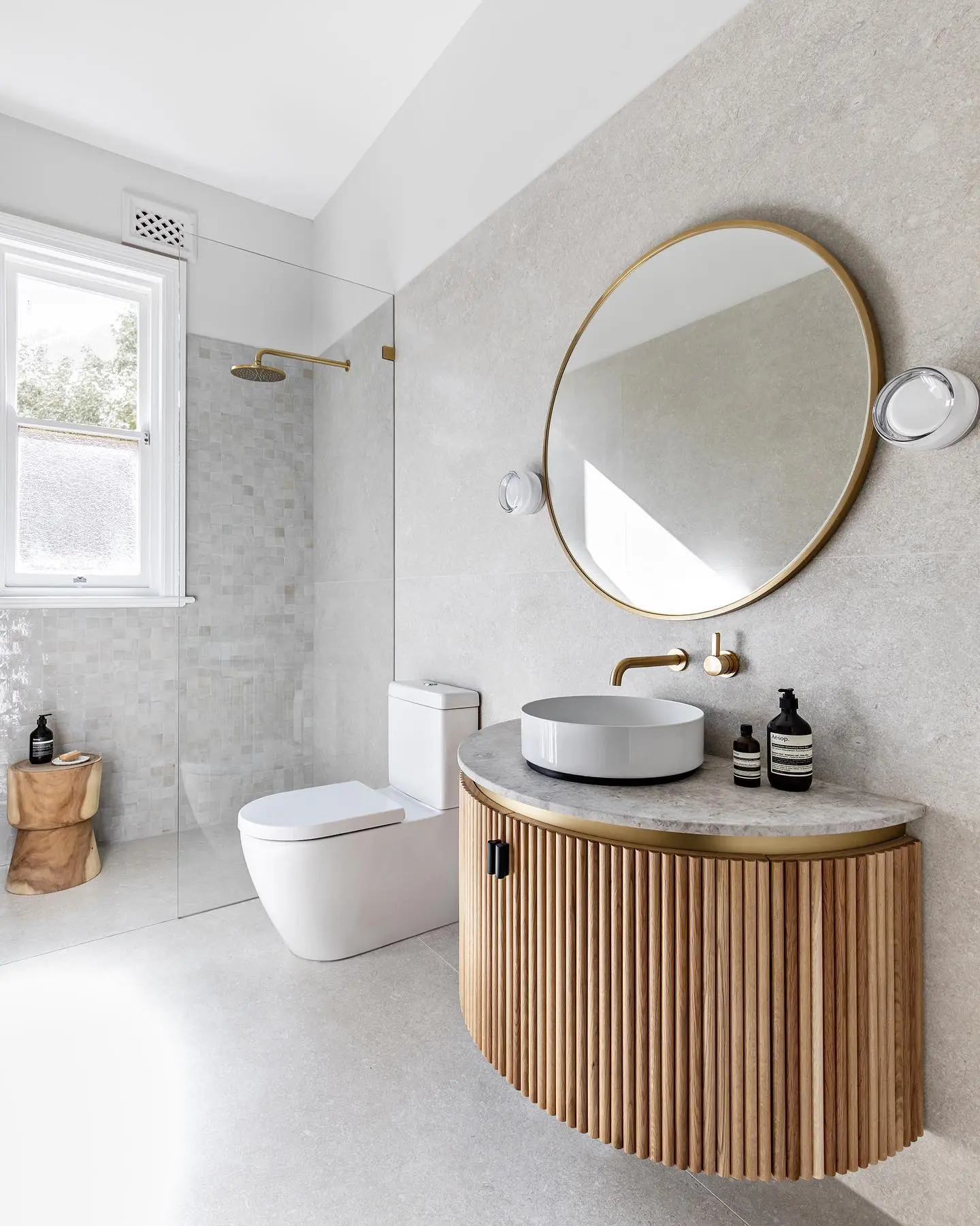 A round, light-wood bathroom vanity with a fluted texture on the cabinet and a white marble top. It holds a white vessel sink and is paired with a gold-framed round mirror and gold fixtures, against a light gray tiled wall.