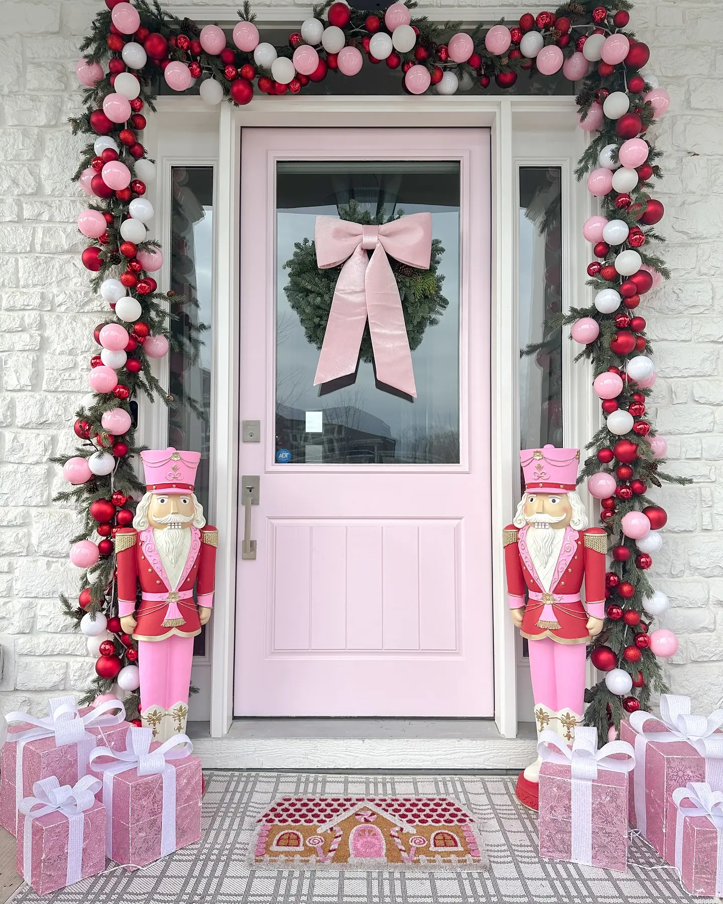 A colorful, non-traditional Christmas porch with a light pink door, surrounded by garland heavily decorated with pink, red, and white ornaments, flanked by two pink and red Nutcracker statues and sparkly pink gift boxes.