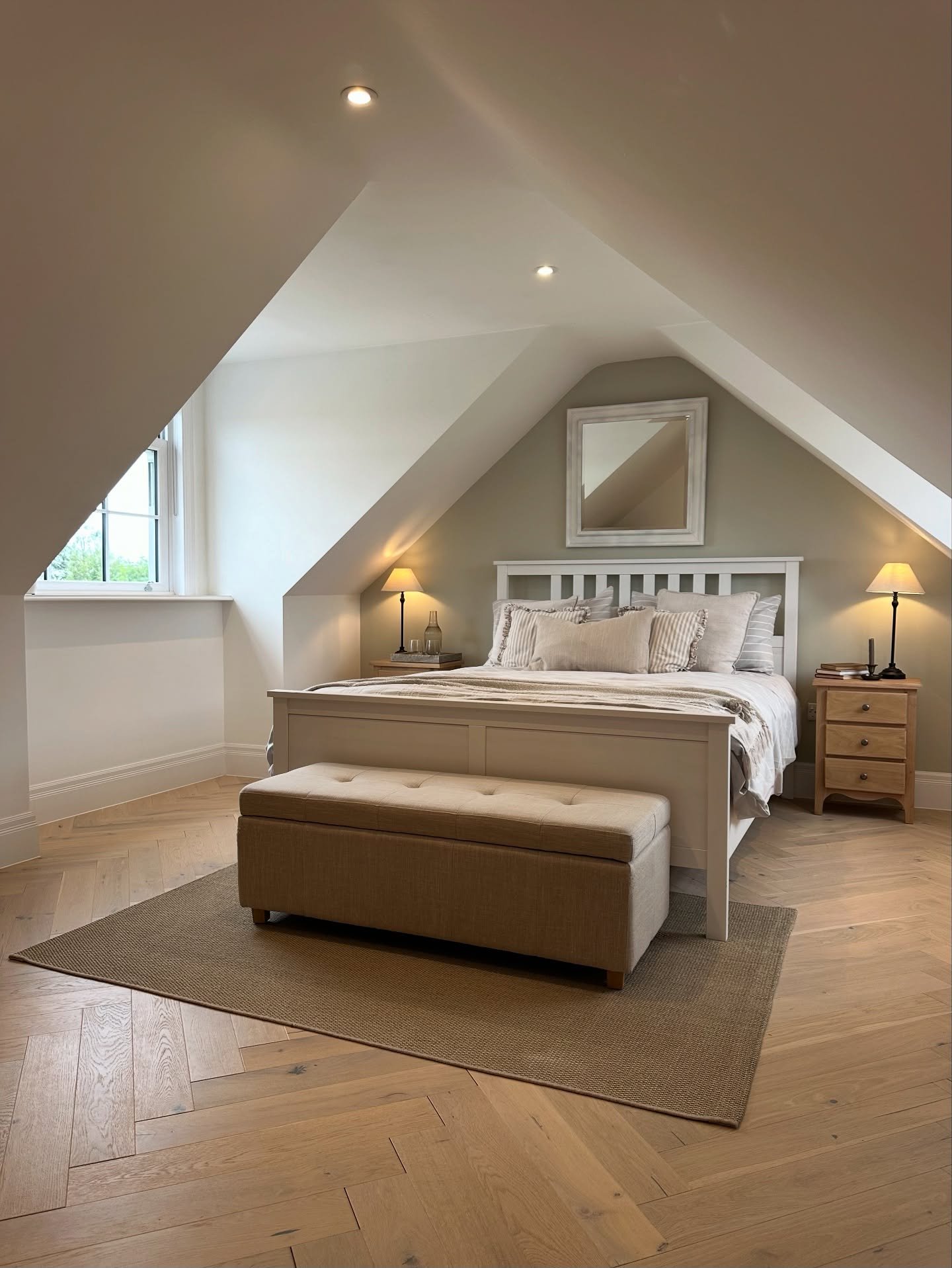 A cozy loft bedroom with a white bed, beige upholstered storage bench, and light wood herringbone floors. The walls are a mix of white and soft sage green.