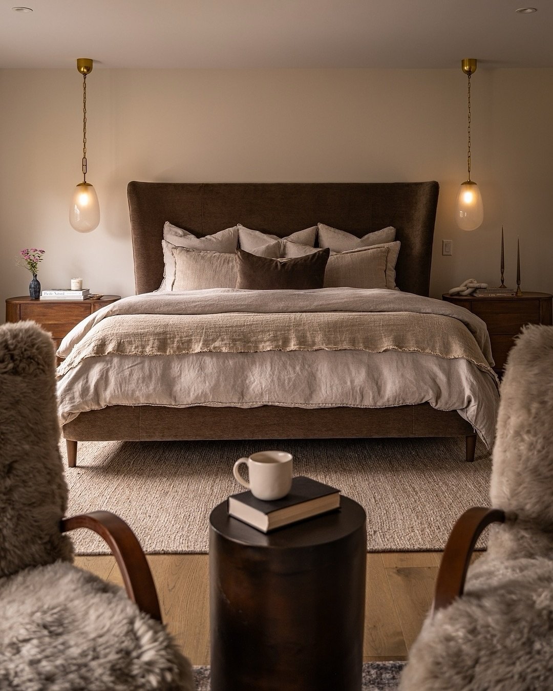 A cozy bedroom with a dark brown upholstered bed, warm lighting from hanging pendants, and two fluffy, textured armchairs in the foreground.