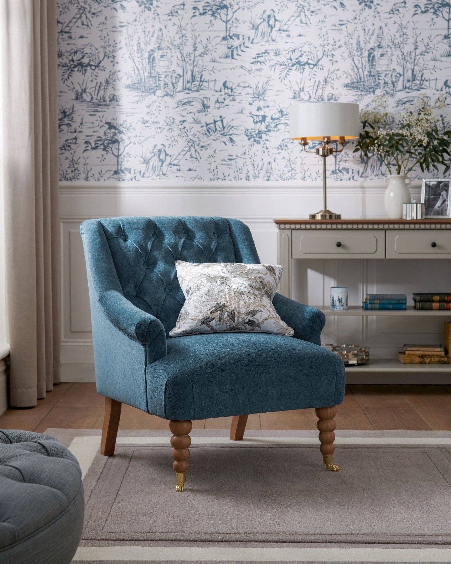 A button-tufted blue-green velvet armchair with turned wooden legs sits on a rug in a traditional-style bedroom.