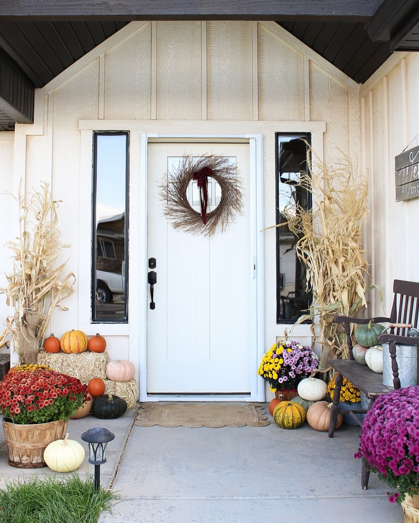 A rustic farmhouse porch decorated for fall with corn stalks, a mix of pumpkins, mums in woven baskets, and a simple grapevine wreath on the white door.