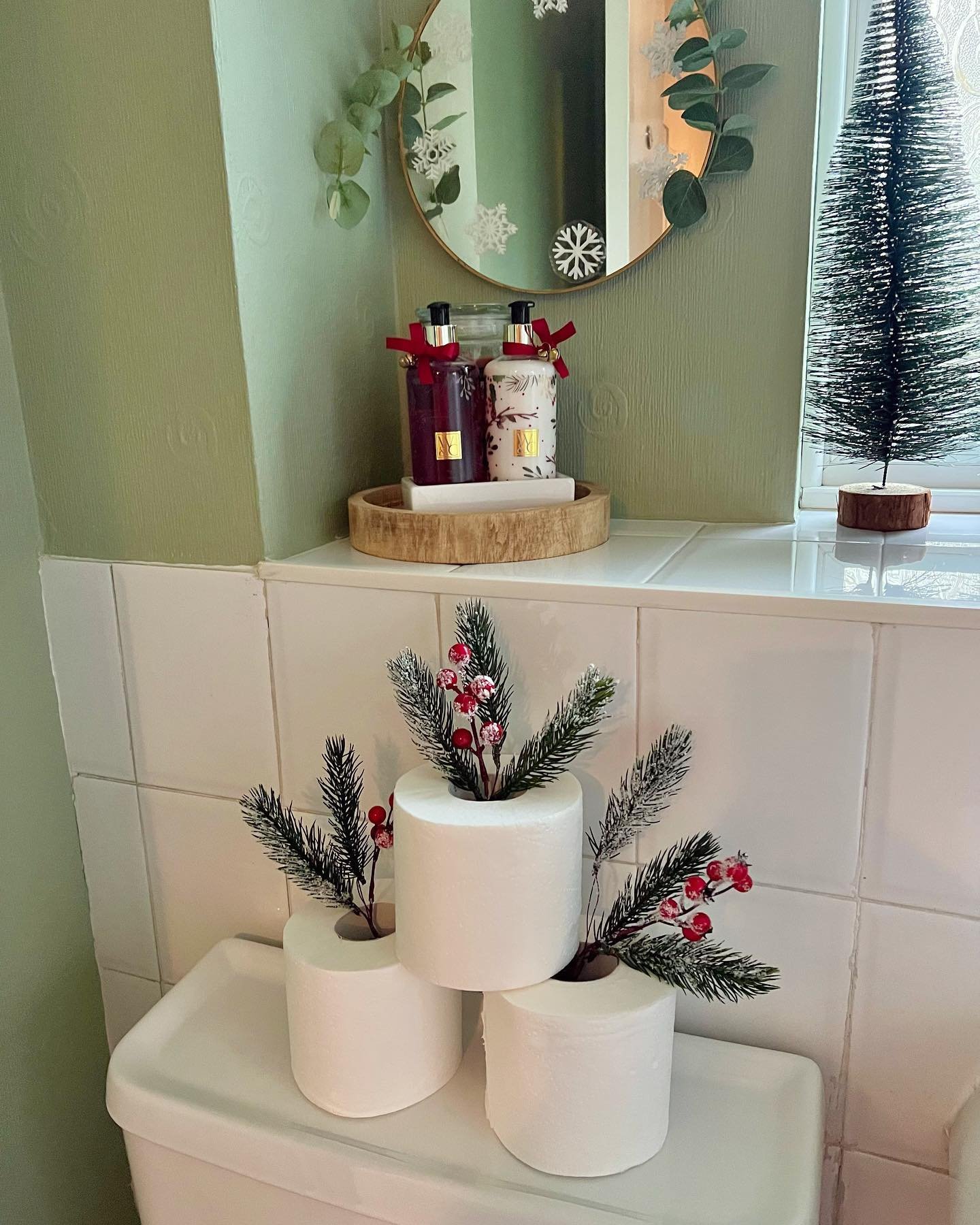 Toilet paper rolls stacked on a toilet tank with faux pine branches and red berries tucked into the center of the rolls, next to a wood tray of holiday soap.