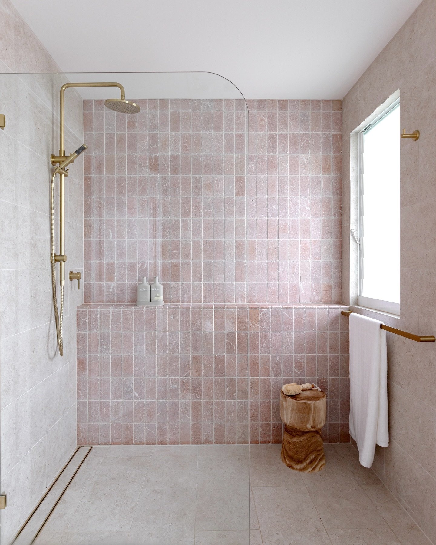 A modern walk-in shower with a wall of square, soft pink-colored tiles with varied, mottled textures and a natural wood stool.