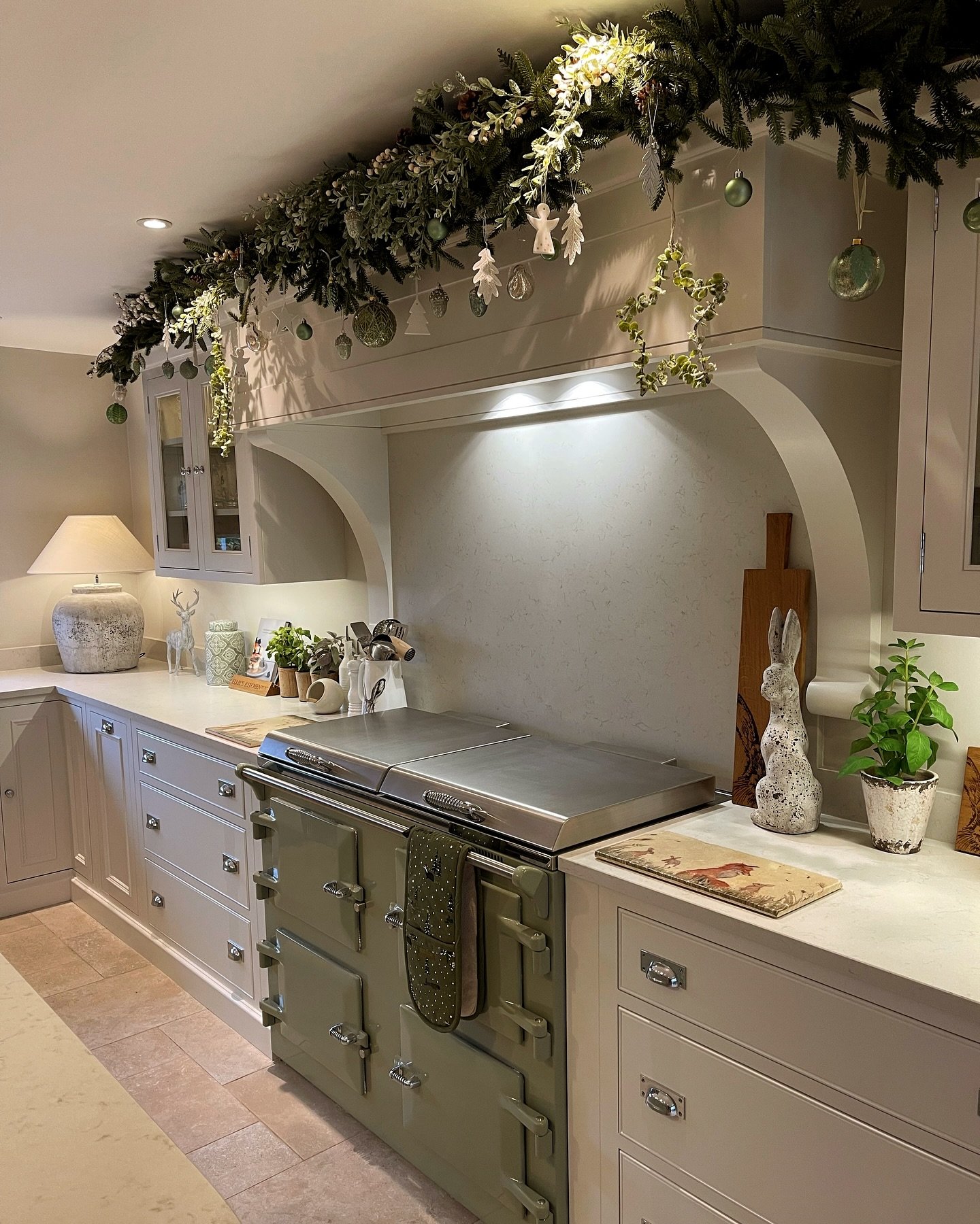 A neutral kitchen featuring an olive green AGA range oven and a custom range hood heavily draped with a luxurious pine garland mixed with pale green and white accents and delicate hanging ornaments.