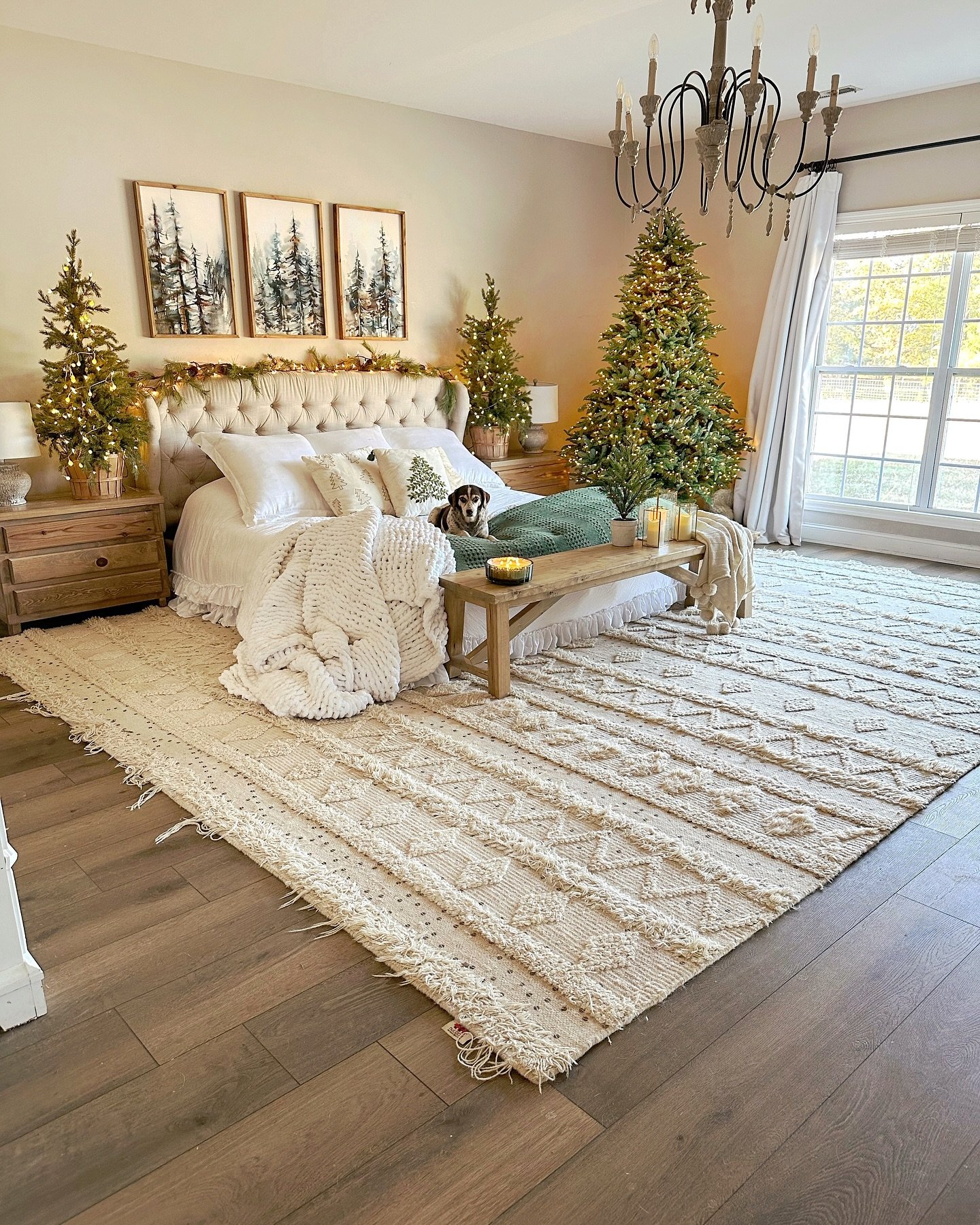 A spacious primary bedroom with a large textured rug, featuring a bed with white bedding, three framed forest prints above the headboard, and three illuminated Christmas trees of varying sizes.