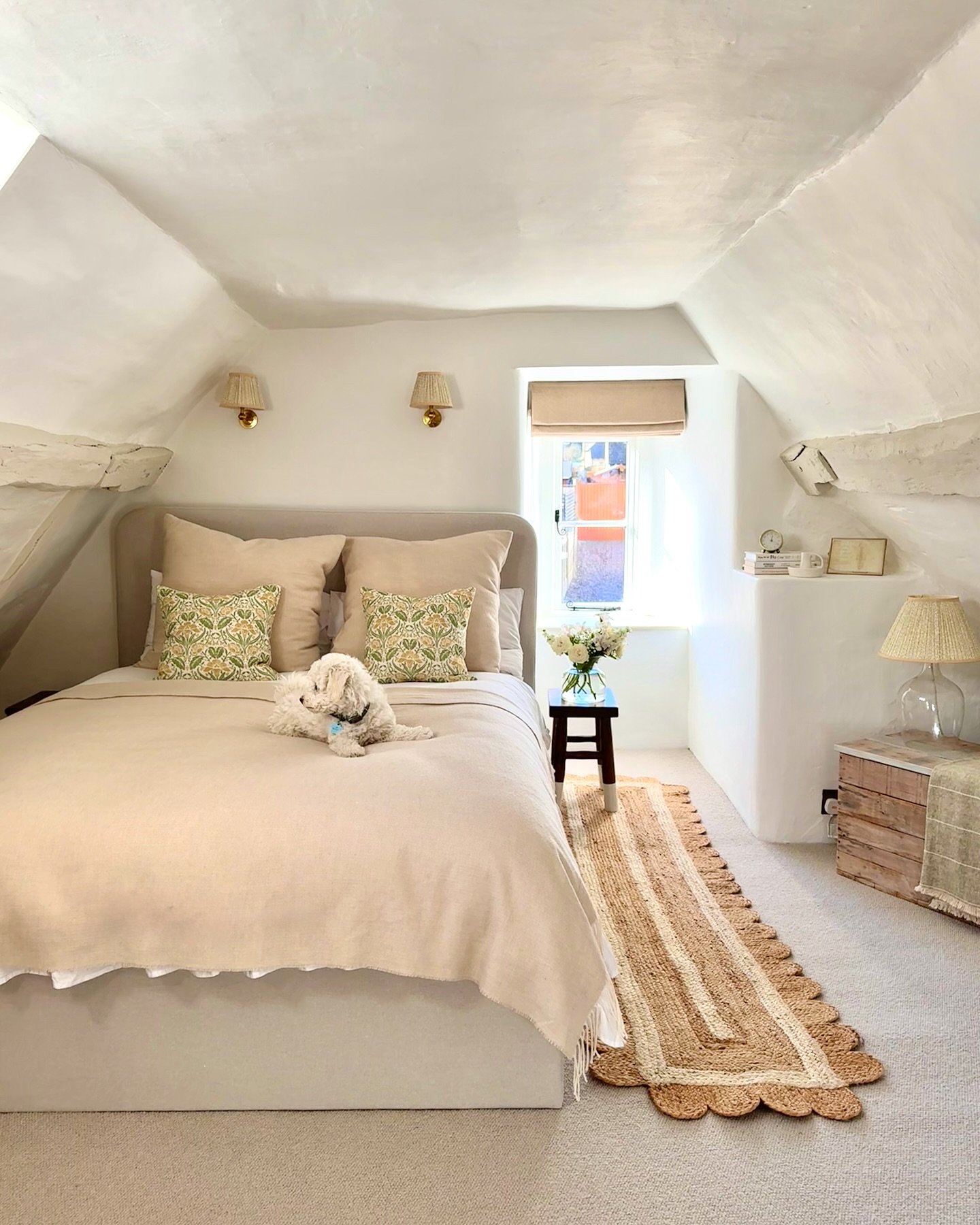A cozy attic bedroom with a sloped ceiling, a bed with neutral bedding and floral pillows, a jute rug, and a white fluffy dog resting on the bed.