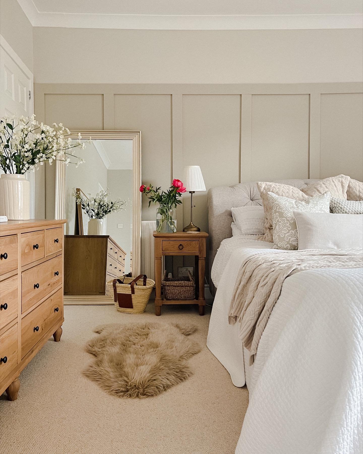 A classic, gold-framed rectangular mirror leans on a wall in a neutral-toned bedroom, reflecting a wooden dresser and a large vase of flowers.