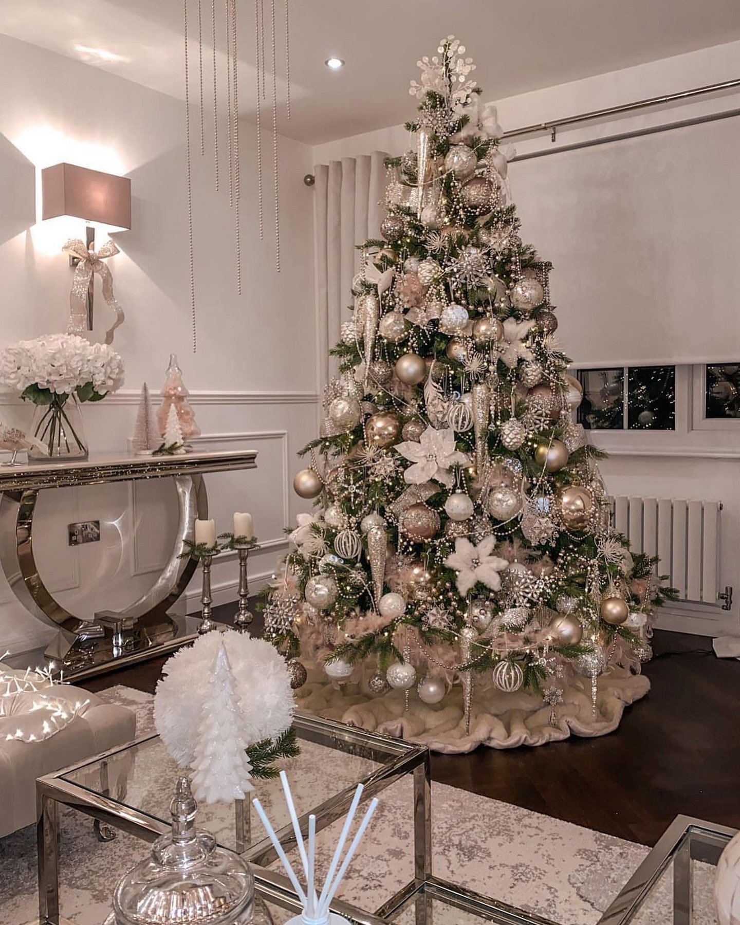 A heavily decorated Christmas tree in a romantic style, featuring pale pink, rose gold, and cream ornaments, dangling beaded garland, and large floral accents.
