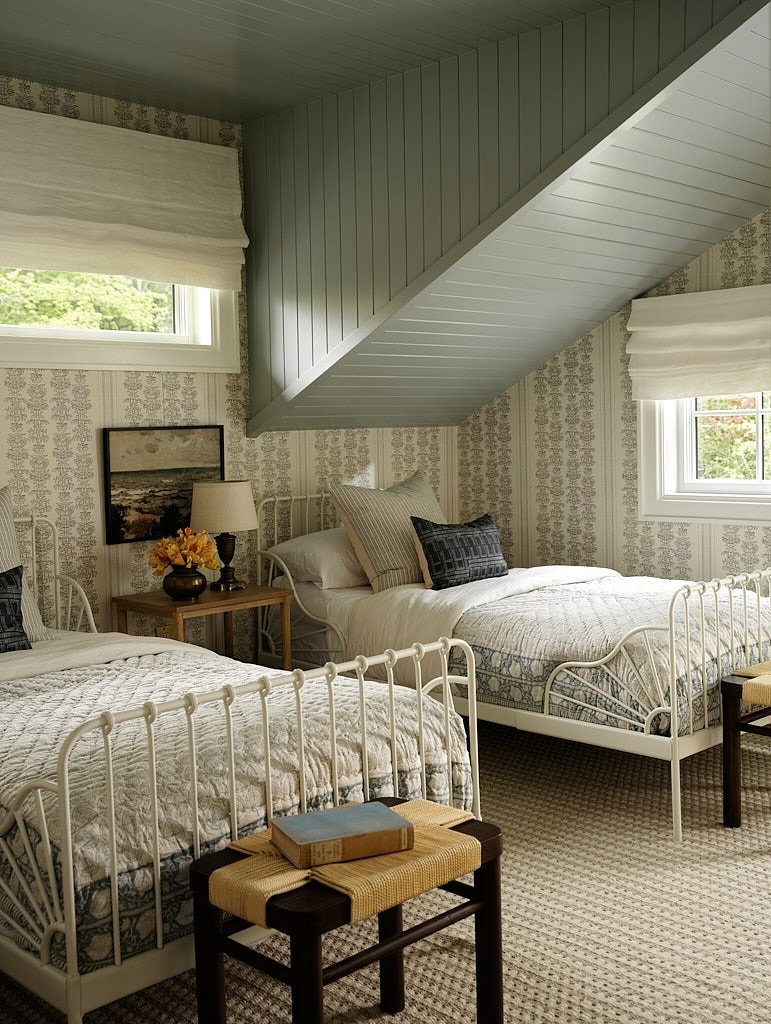 A cozy twin bedroom with two white metal beds and blue patterned quilts, featuring a unique angled ceiling and cottage-style decor.