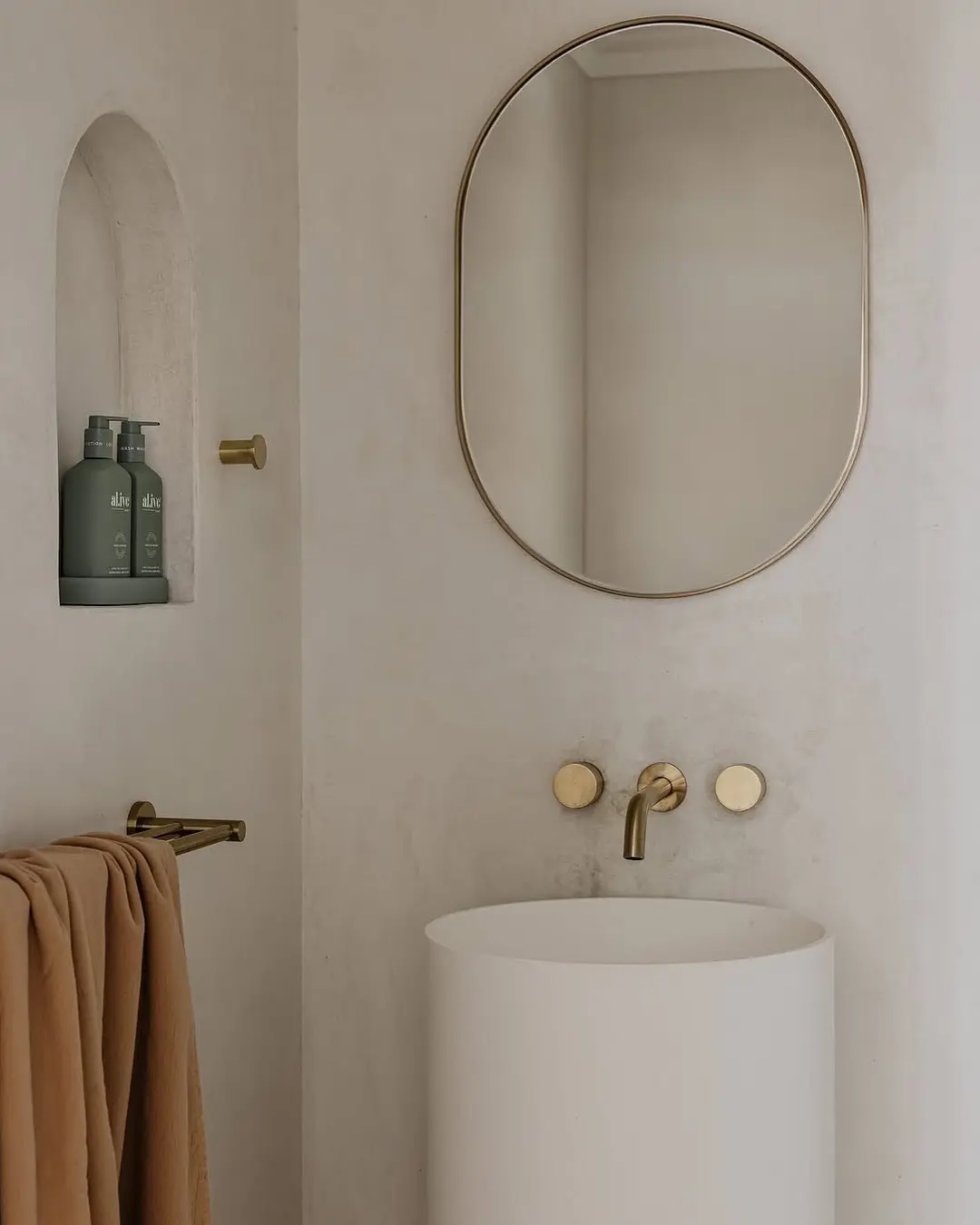 A minimalist bathroom featuring a pill-shaped mirror with a thin brass frame above a round, white pedestal sink.