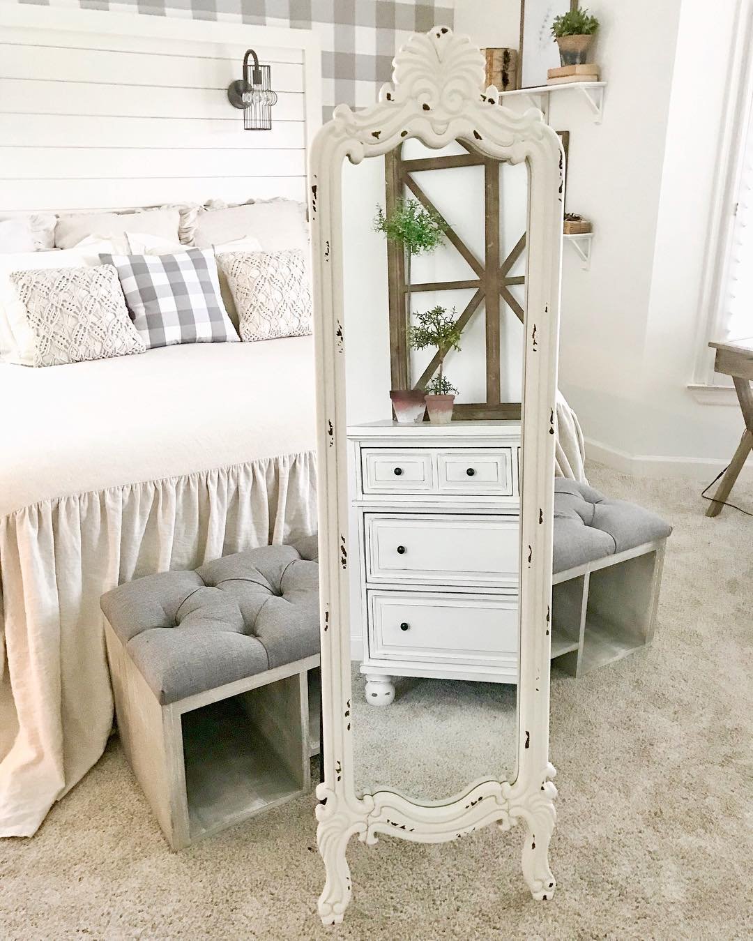 A vintage-style, freestanding mirror with an ornate, distressed white frame stands in a rustic-themed bedroom, reflecting a white dresser and tufted bench.