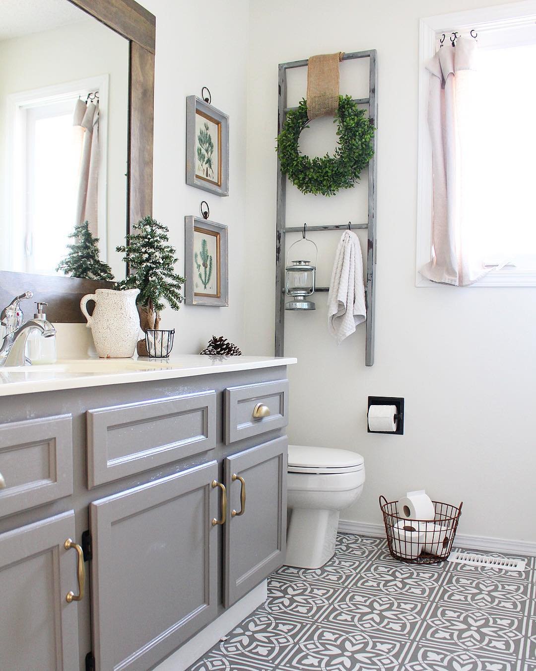A white and grey bathroom with a decorative grey ladder holding a boxwood wreath and a small wire basket of toilet paper on the floor.