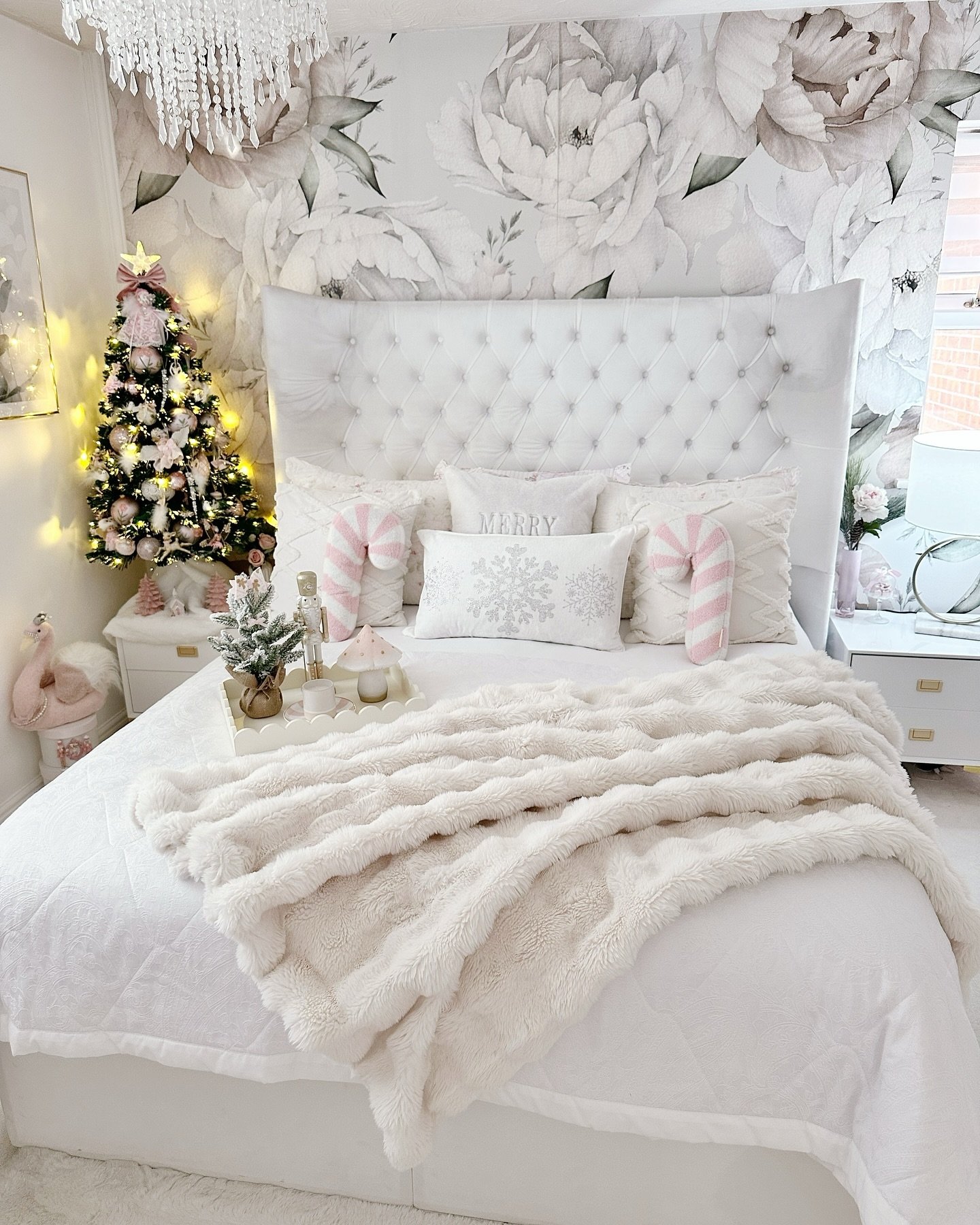 A luxurious white bedroom with a floral accent wall and crystal chandelier, featuring a bed dressed in white bedding, a pale pink Christmas tree, and pink candy cane accent pillows.