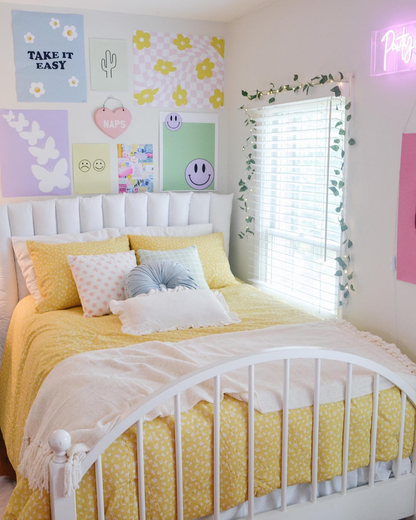 A bright and colorful bedroom with a white metal bed, a yellow floral comforter, a gallery wall of pastel art prints, and a pink neon sign.