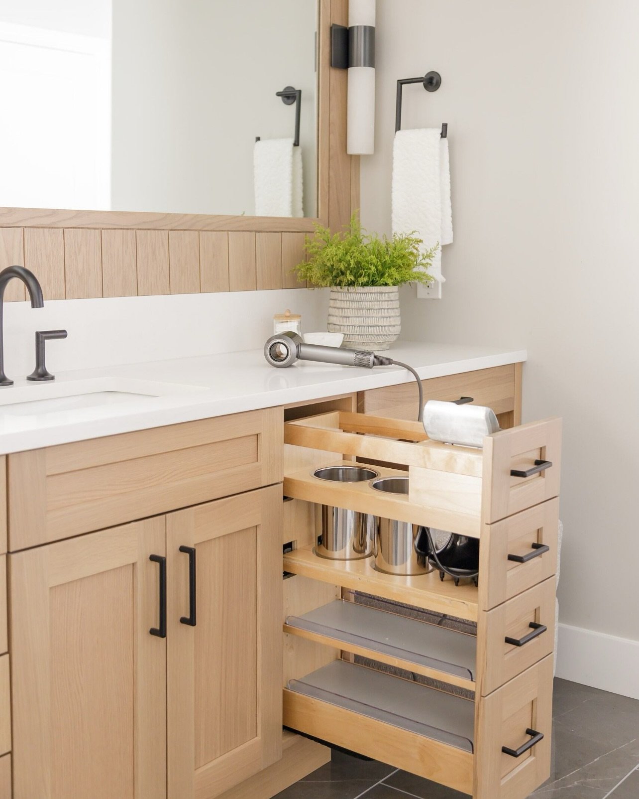 Light natural wood bathroom vanity with a specialized narrow pull-out tower caddy featuring built-in metal cylinders and shelves for storing a hairdryer and hot tools.