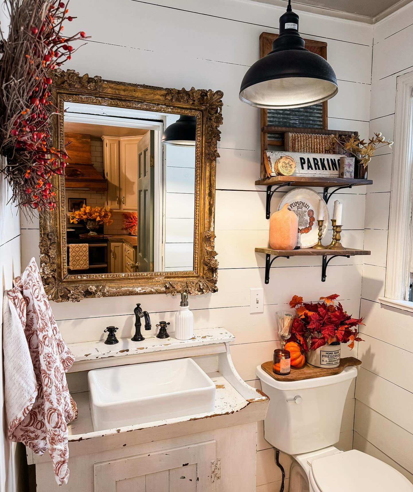 A small bathroom with shiplap walls, a vintage gold mirror, and shelves decorated with fall items like a wreath and pumpkins.