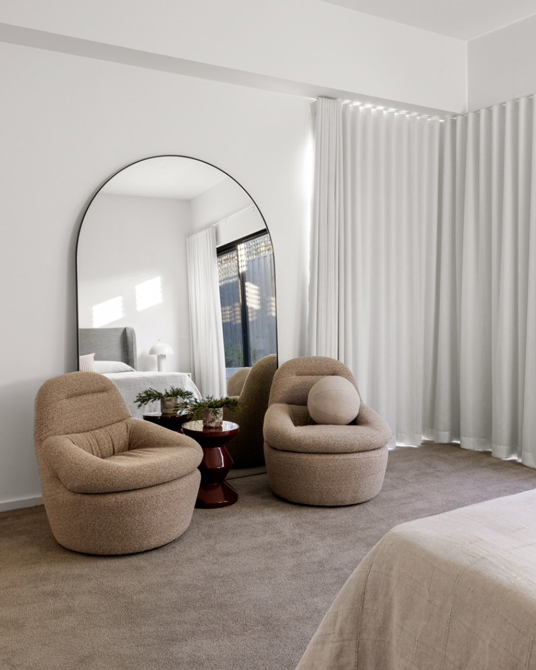 Two modern, rounded armchairs in a textured beige fabric sit in front of a large arched mirror.