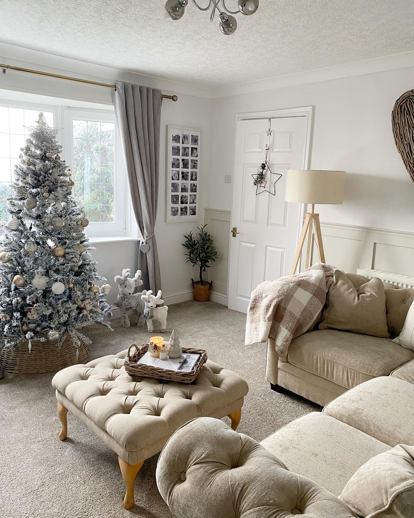 Cozy living room featuring a beige sofa, tufted ottoman, and a silver flocked Christmas tree next to a window.