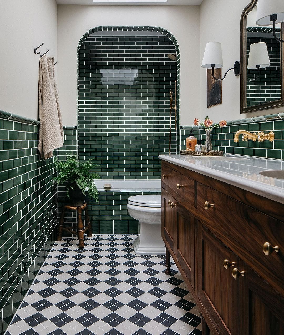 A traditional bathroom with dark green rectangular subway tiles on the lower walls and in an arched shower niche, paired with a black and white diamond-patterned floor.