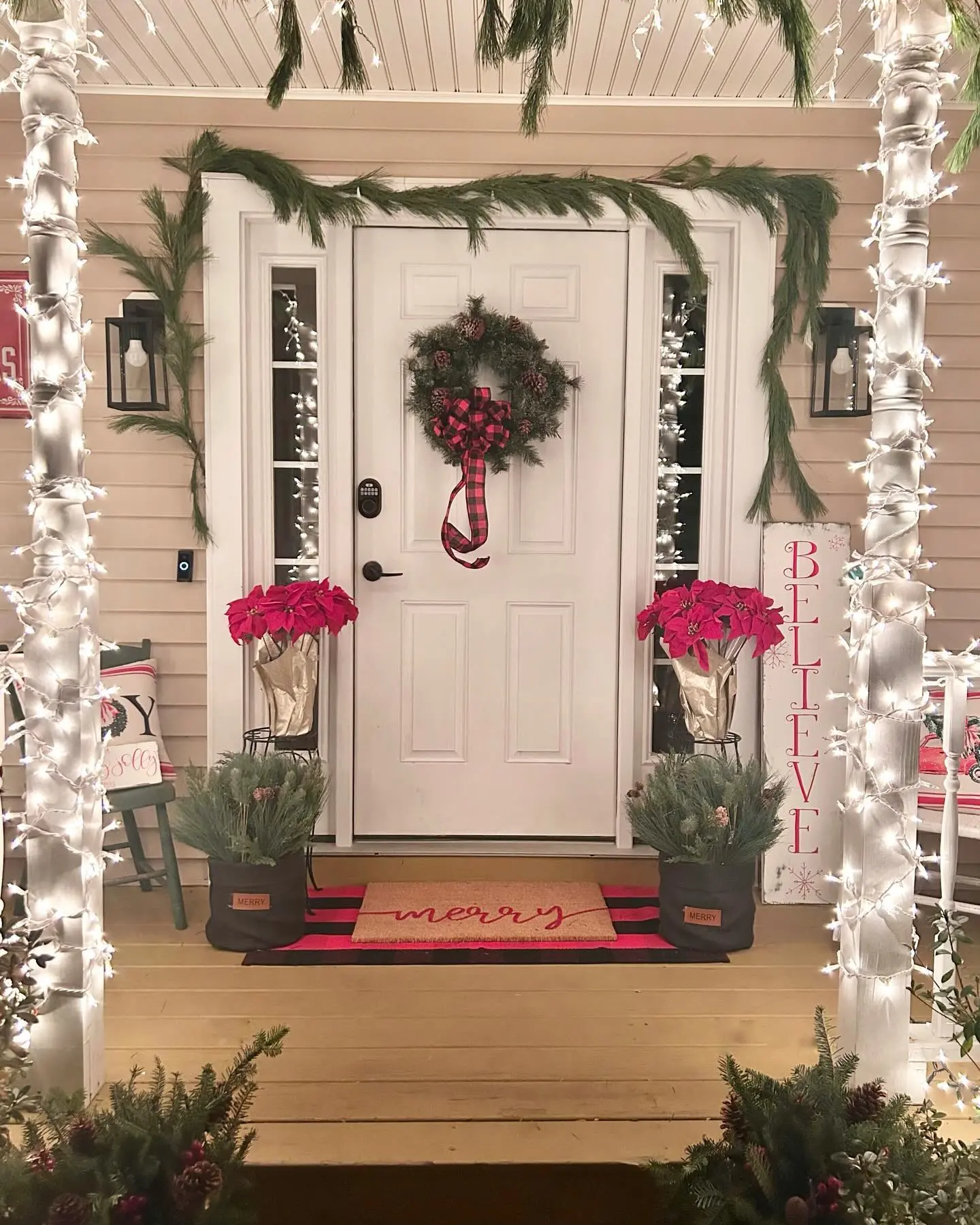 A cozy Christmas porch glowing with pure white string lights wrapped around the pillars, featuring a white door with a wreath tied with a red and black buffalo plaid ribbon, and two planters of red poinsettias.