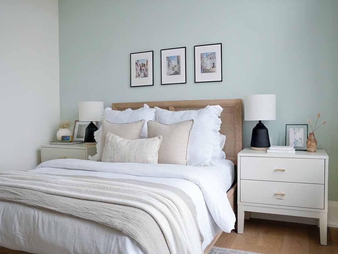 A matching pair of white, two-drawer nightstands with gold bar pulls, placed on either side of a bed with a wooden headboard. The nightstands are styled with black lamps, a digital clock, a vase of dried flowers, and framed pictures.