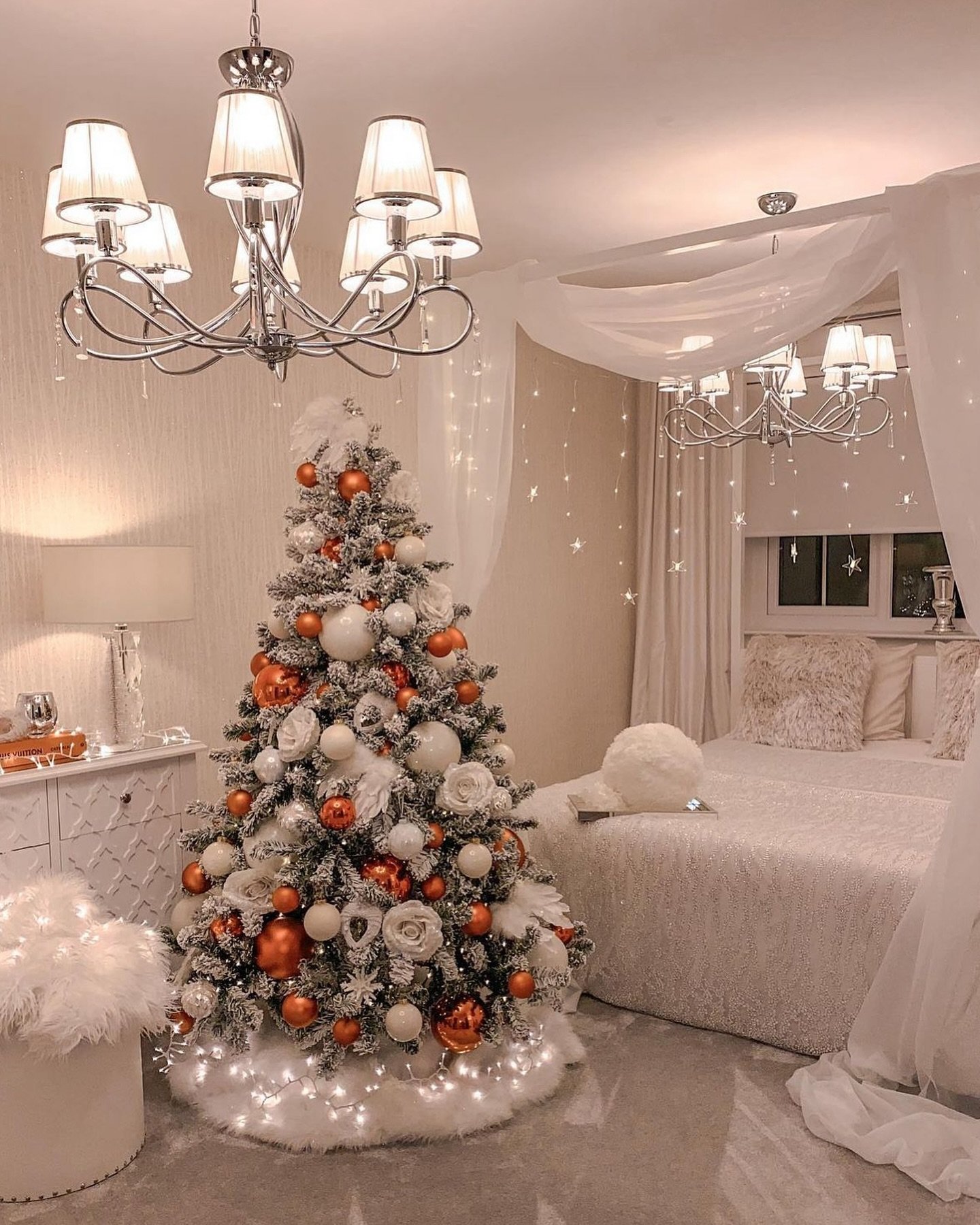 A flocked Christmas tree decorated with large white flowers, glossy white ornaments, and bright copper baubles, set in a cozy, elegant bedroom.